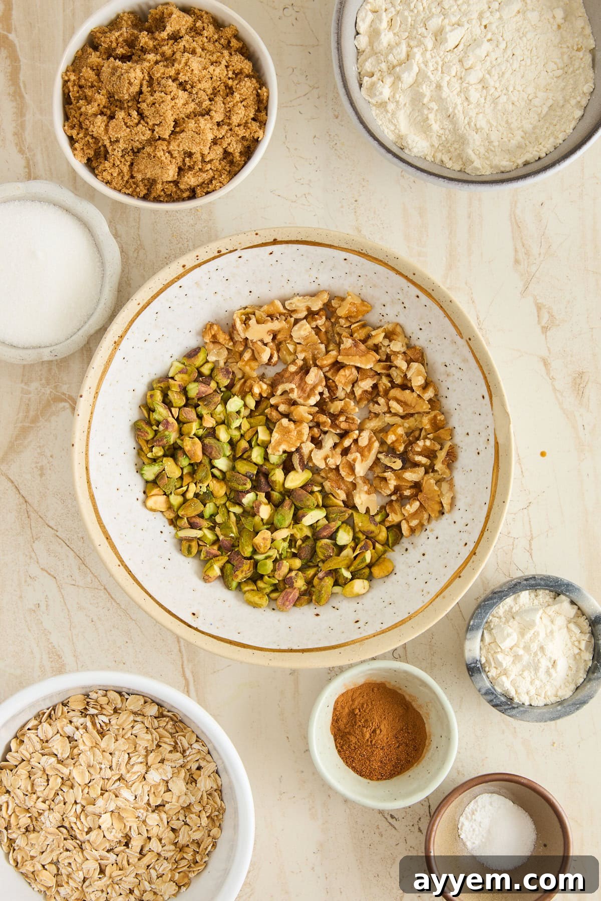 A variety of baking ingredients in bowls on a light surface, including walnuts, pistachios, brown sugar, flour, oats, cinnamon, salt, and granulated sugar, ready for making Baklava Apple Crisp.