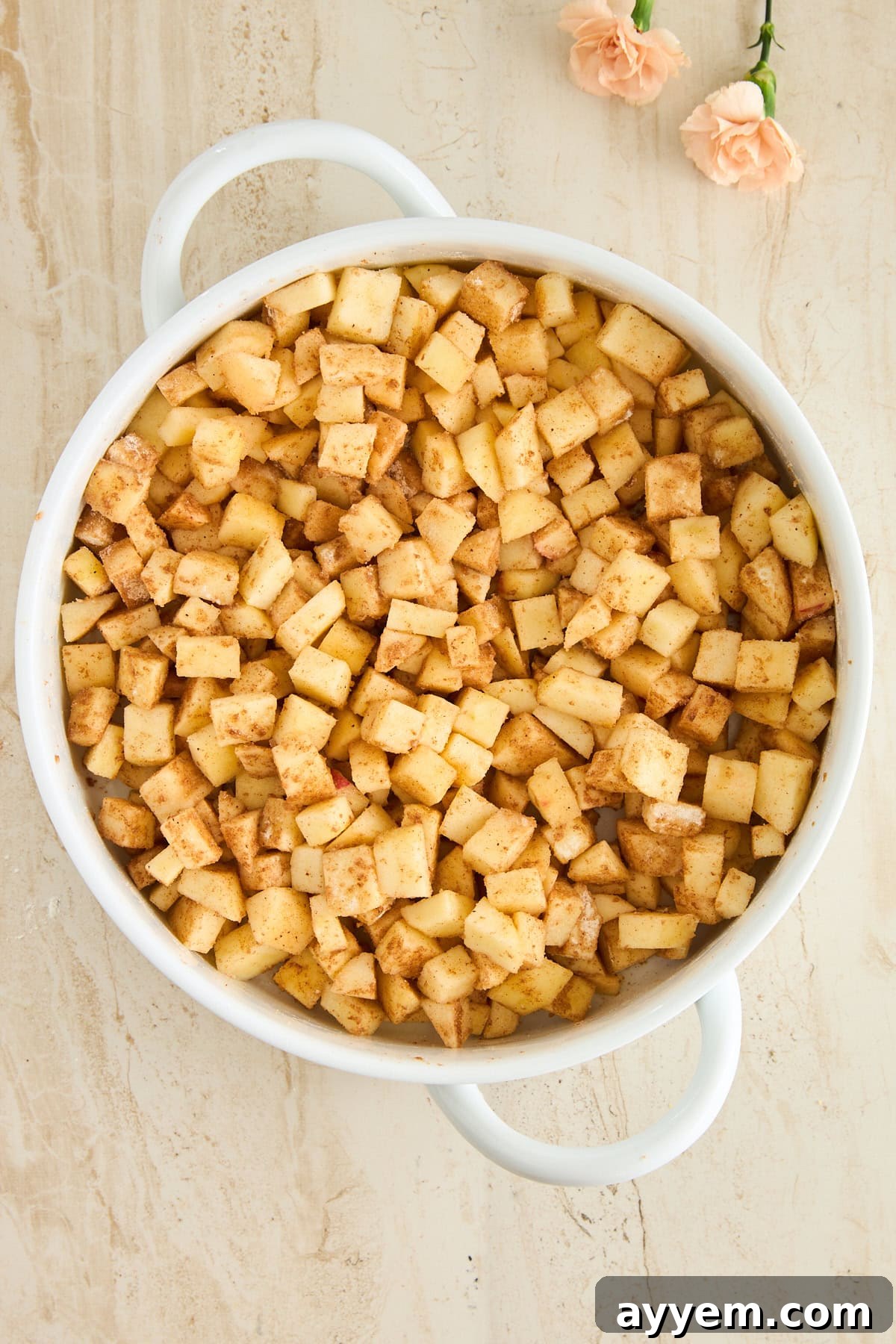A white pot filled with perfectly diced Granny Smith apples coated in cinnamon and sugar sits on a light beige surface, with two light pink flowers placed nearby, showcasing the filling ingredients for Baklava Apple Crisp.