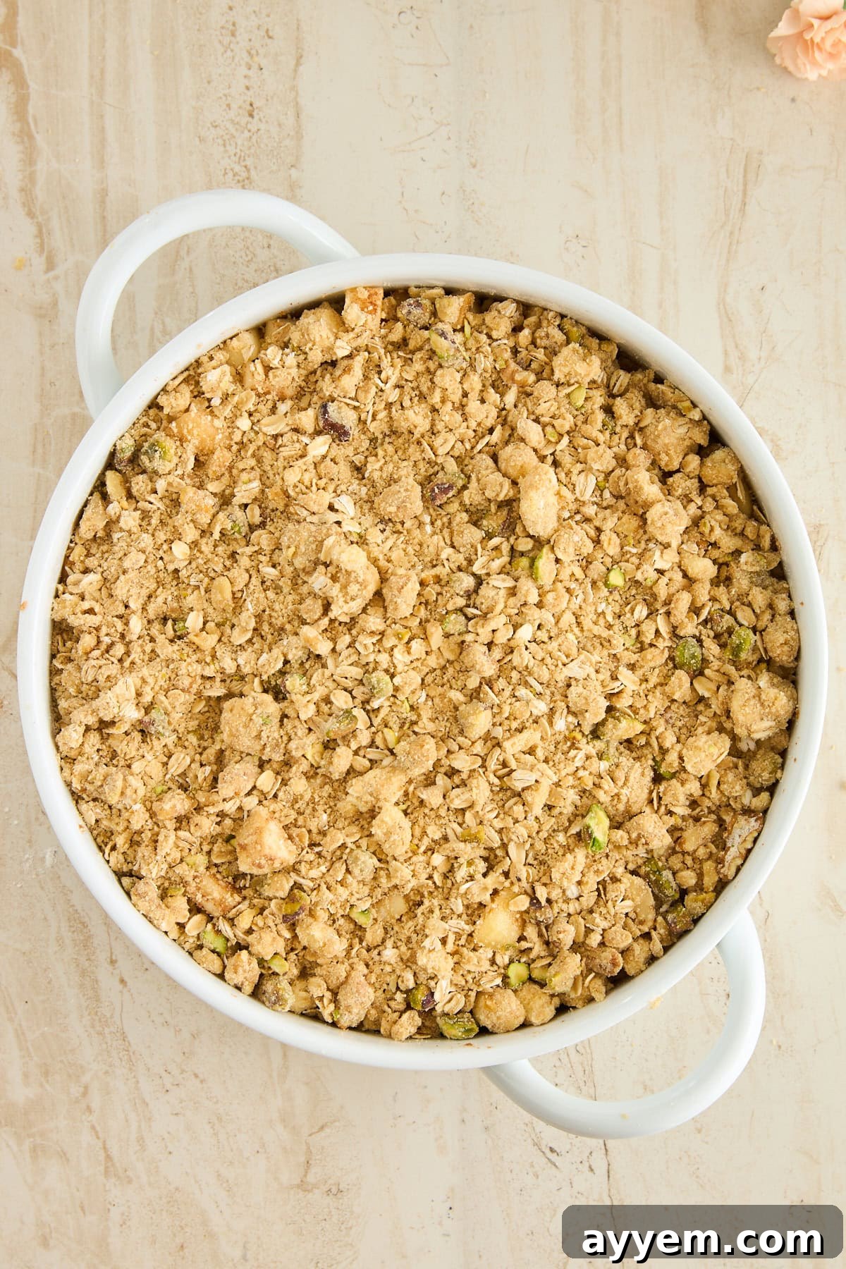 A round white baking dish with handles, filled with a golden-brown oat and nut crumble topping before baking, set on a light beige textured surface. This is the Baklava-inspired streusel.