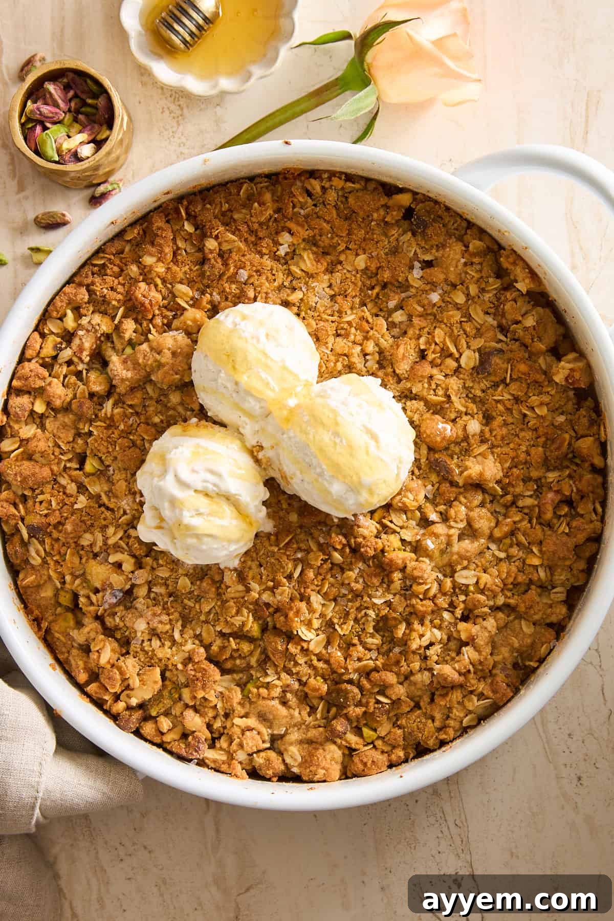 A round white dish filled with golden-brown Baklava Apple Crisp, generously topped with three scoops of creamy vanilla ice cream drizzled with honey. A small bowl of pistachios, a wooden honey dipper, and a pale rose rest nearby on a light surface, highlighting the elegant presentation.