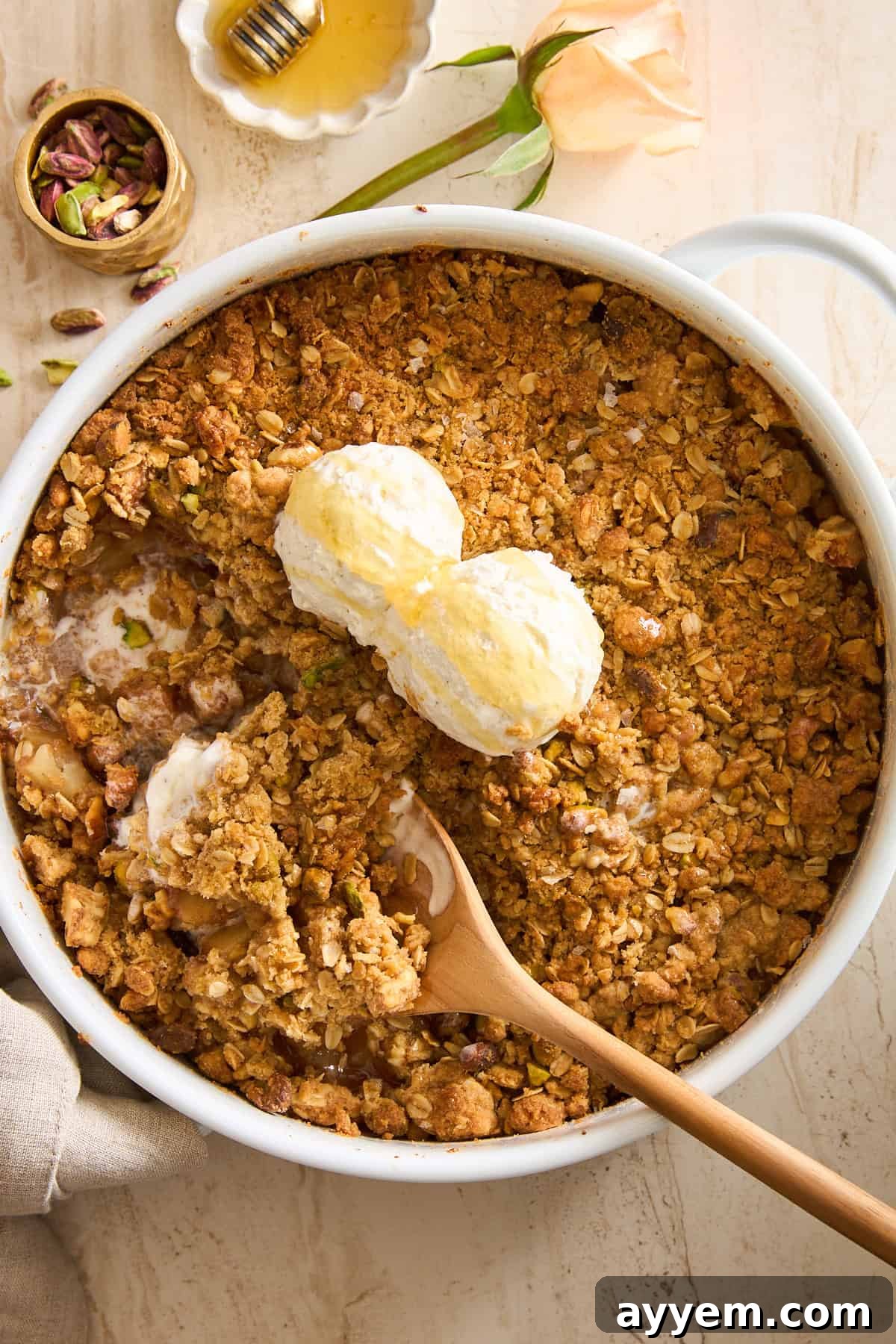 A baked fruit crisp topped with two scoops of vanilla ice cream sits in a round white dish. A wooden spoon is resting in the dish, and some ice cream is melting into the golden, crumbly topping, ready to be enjoyed.