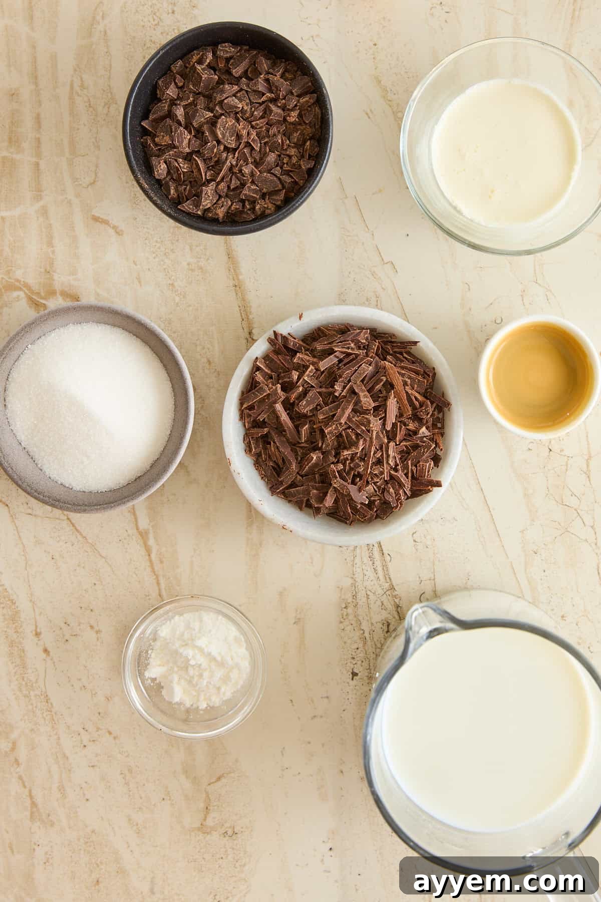 Overhead view of bowls containing chocolate chunks, milk, cream, sugar, a yellow liquid (likely vanilla extract), and a small bowl of cornstarch, arranged on a light-colored marble surface. 