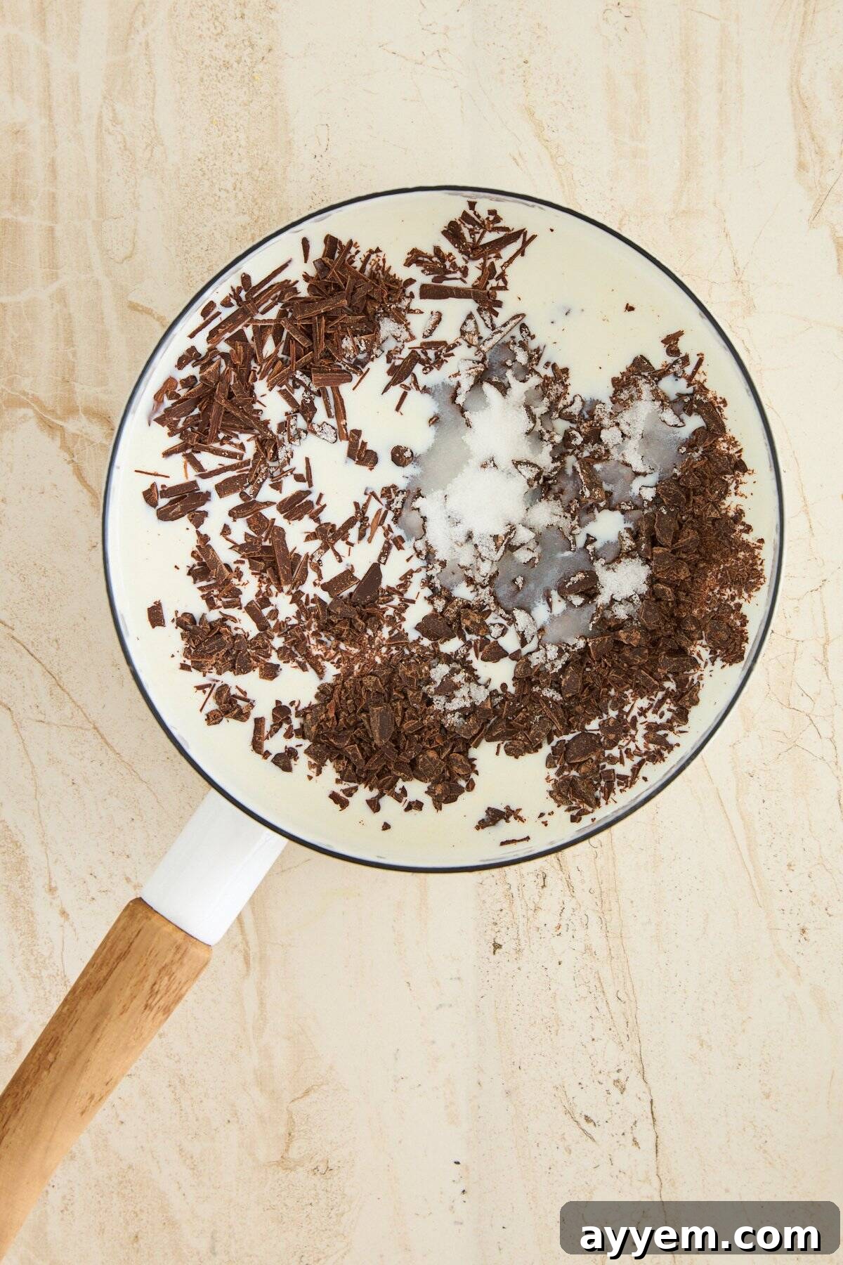 A white saucepan with a wooden handle holds cream, sugar, and chopped dark chocolate, viewed from above on a light beige surface.