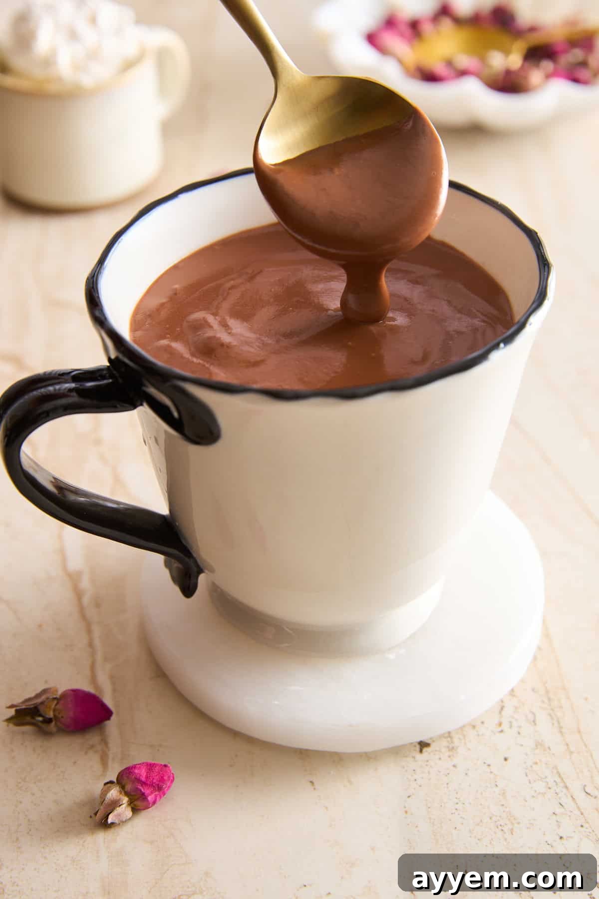 A spoon with thick Italian hot chocolate hovers over a white mug with a black handle, sitting on a white coaster. Rose buds and petals are scattered nearby on a light surface.