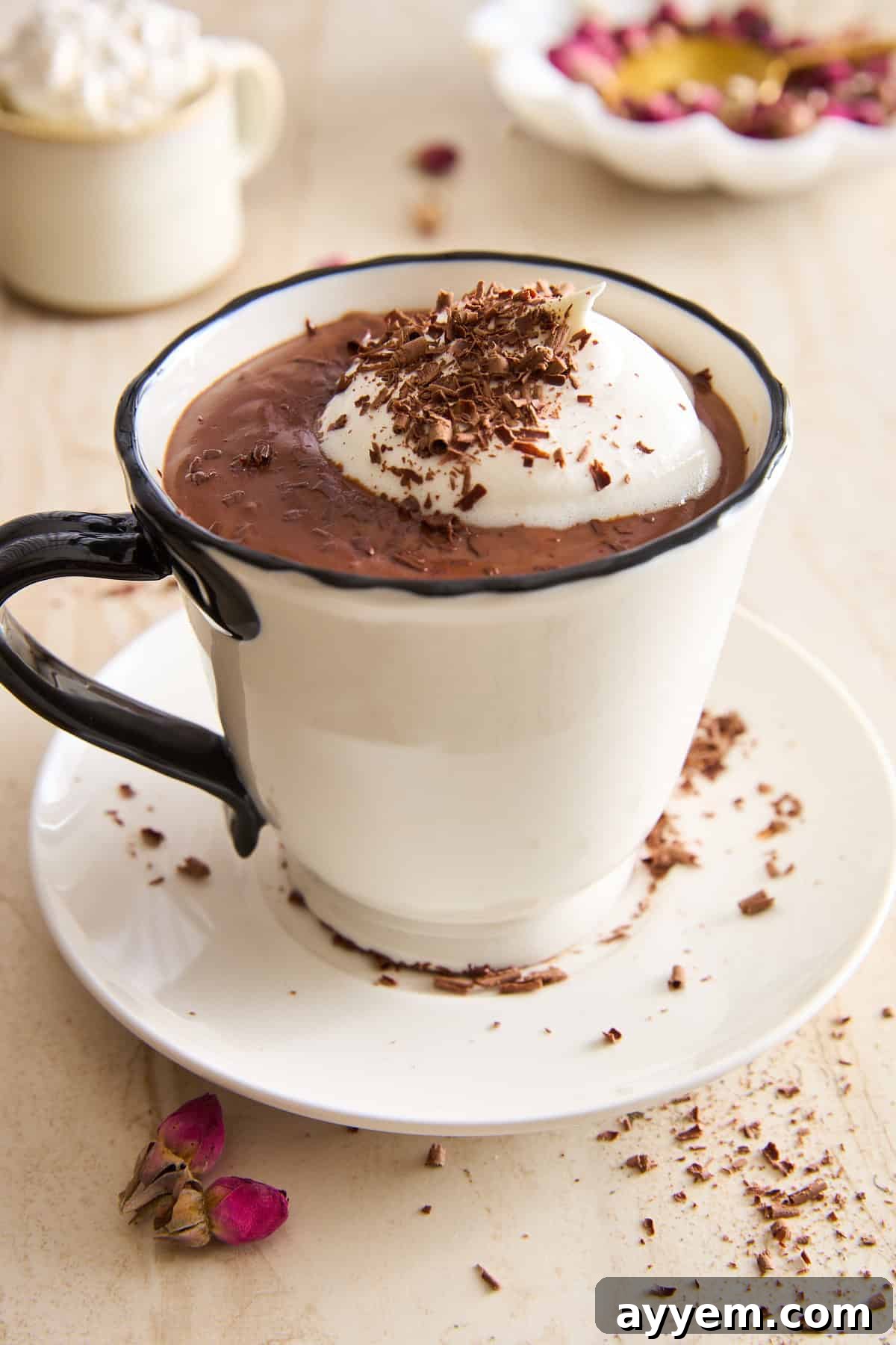A creamy hot chocolate topped with whipped cream and chocolate shavings in a white mug, sitting on a matching saucer, with rose petals and chocolate shavings scattered nearby.