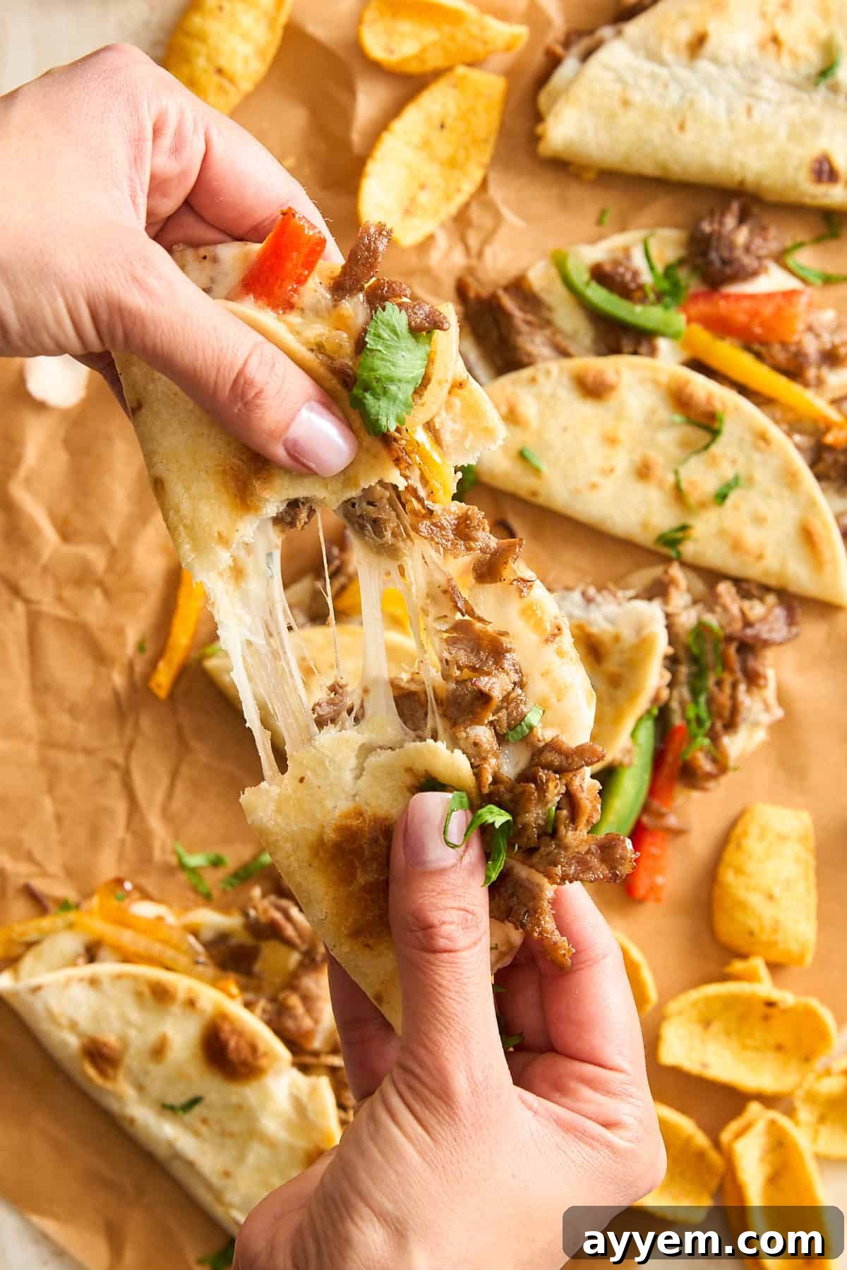 A hand reaching for a cheesy beef Philly cheesesteak taco, with visible peppers and cilantro, against a background of other tacos and corn chips on parchment paper.
