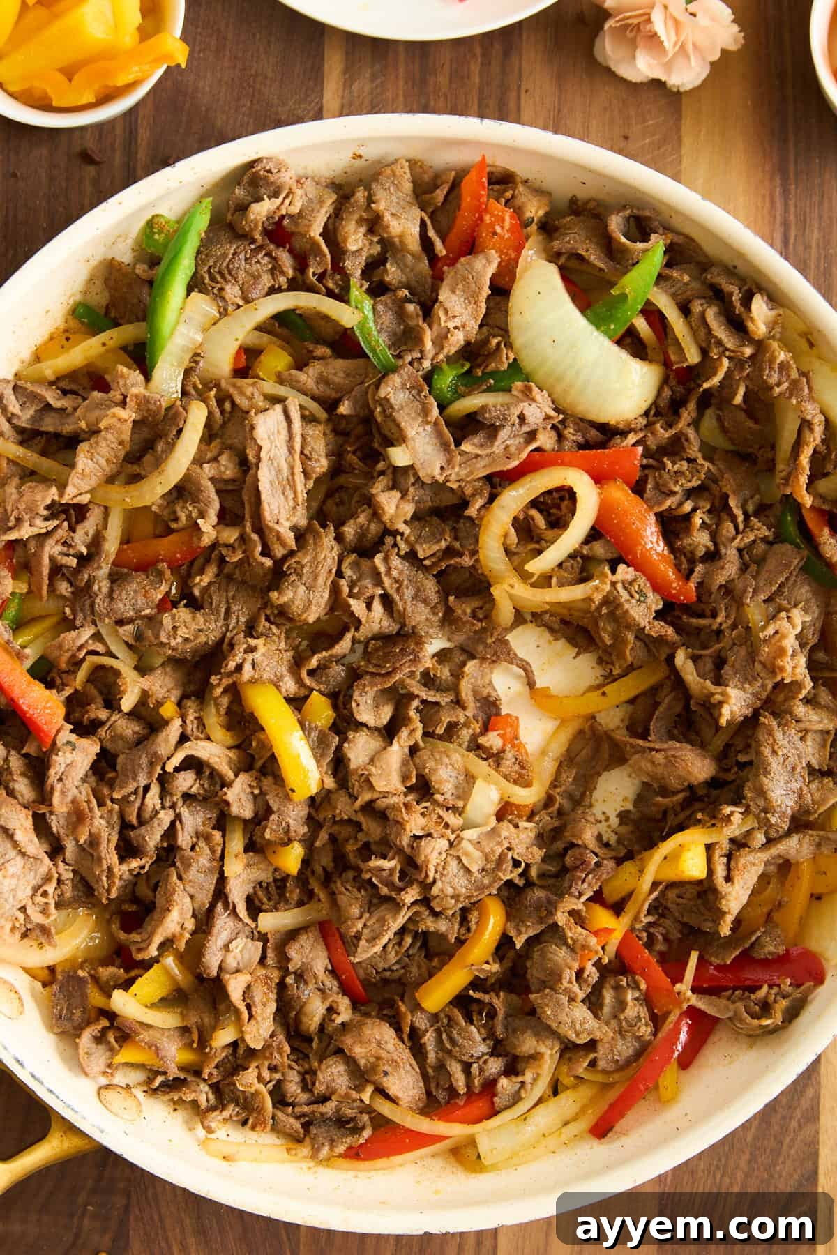A skillet filled with cooked, thinly sliced beef, vibrant yellow and red bell peppers, and tender onions on a wooden surface. The ingredients are beautifully mixed, showcasing the savory filling for the Philly Cheesesteak Pasta Bake.