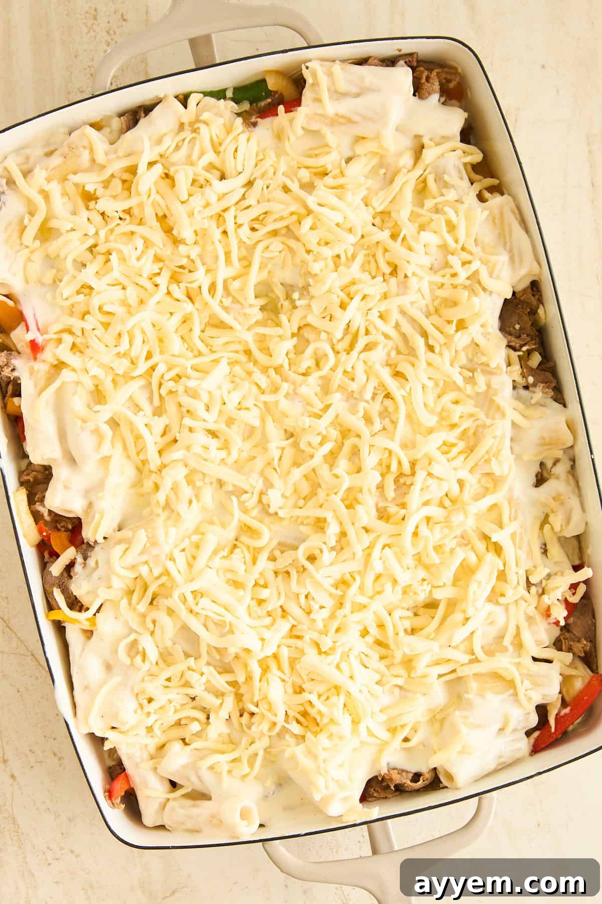 A casserole dish filled with layers of cooked rigatoni, savory sliced vegetables and meat, topped with a creamy white béchamel sauce and a generous amount of shredded cheese, ready to be baked to golden perfection.