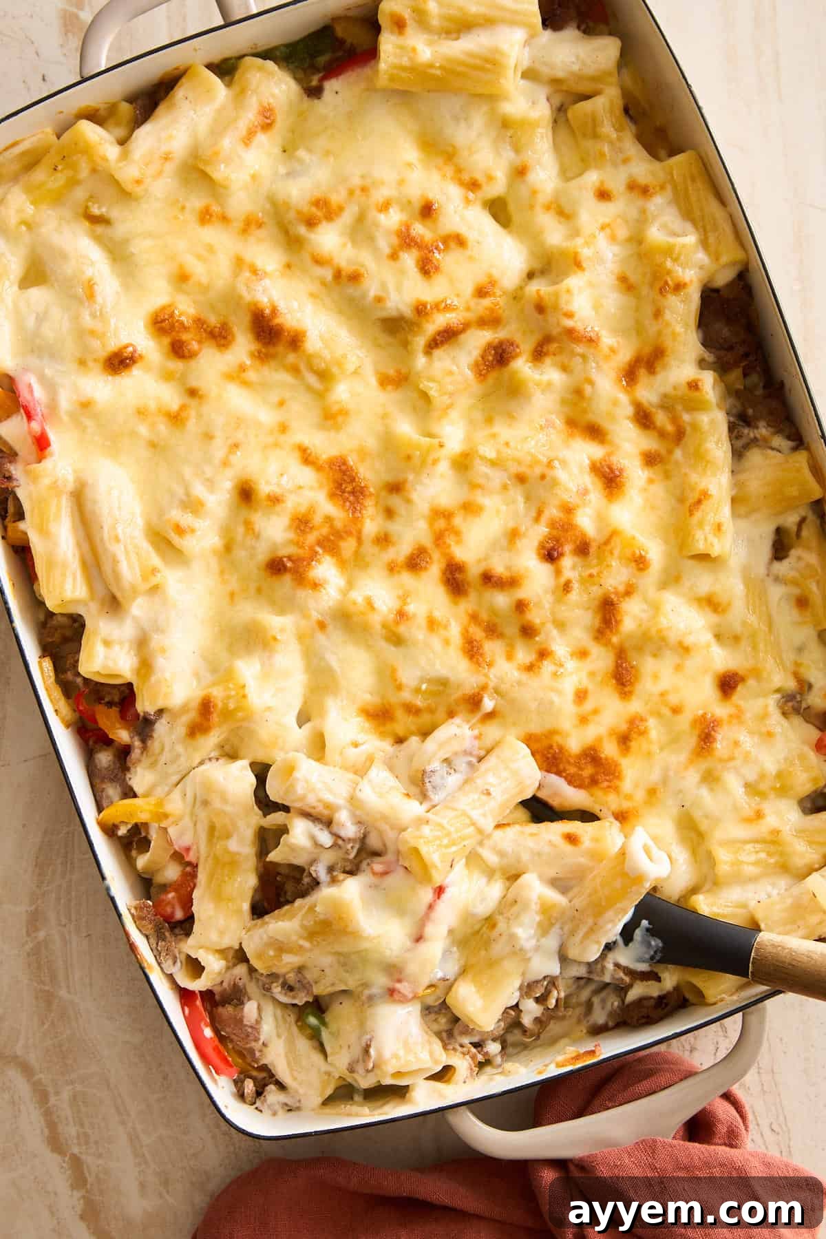 A serving of Philly Cheesesteak Pasta Bake being scooped from a white rectangular baking dish, showing creamy rigatoni, savory steak, and melted cheese, garnished with fresh parsley.