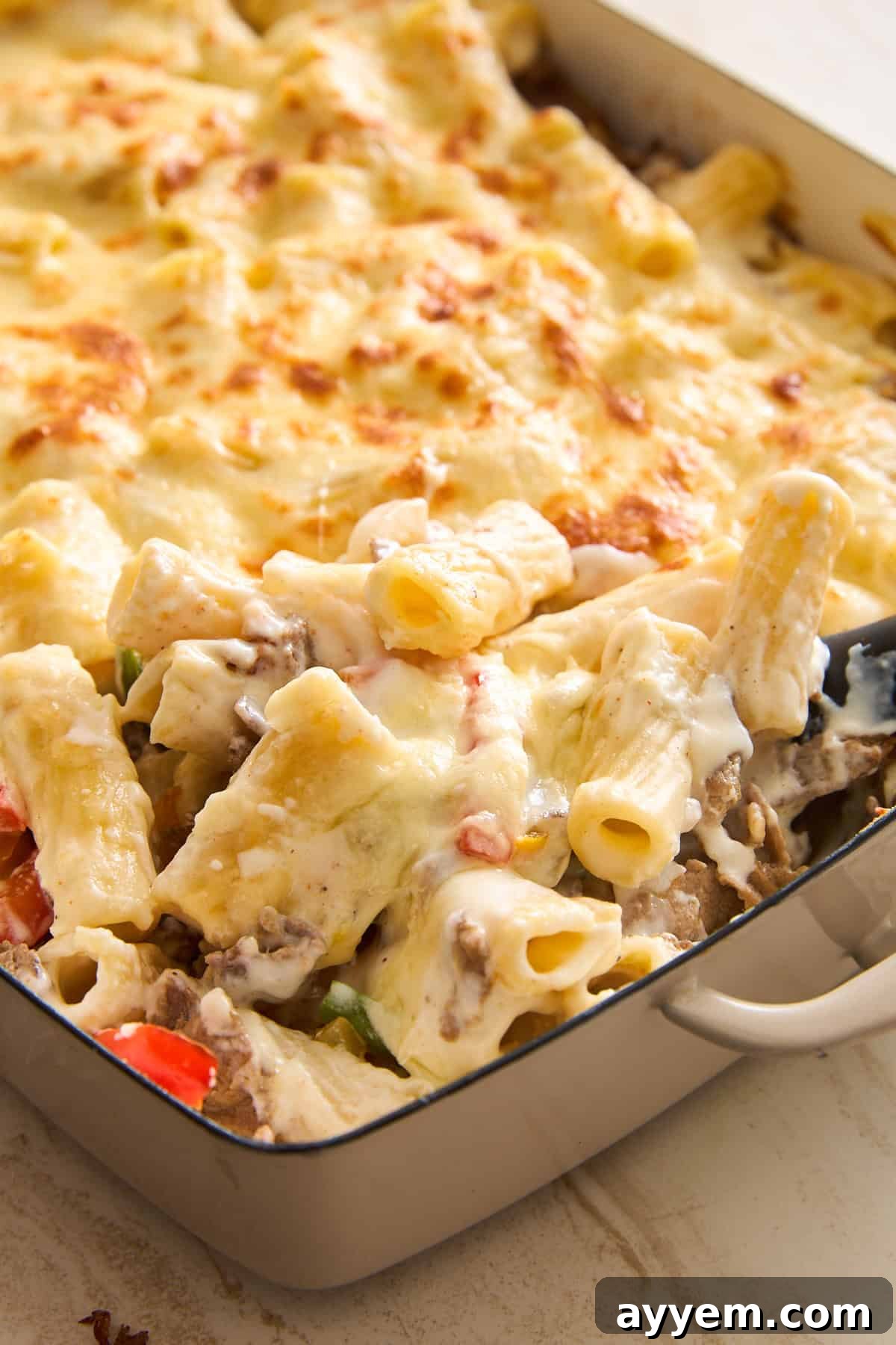 A close-up of a baked pasta casserole in a rectangular dish, featuring rigatoni pasta, melted cheese on top, and visible pieces of vegetables and meat in a creamy sauce. A serving is being scooped out.