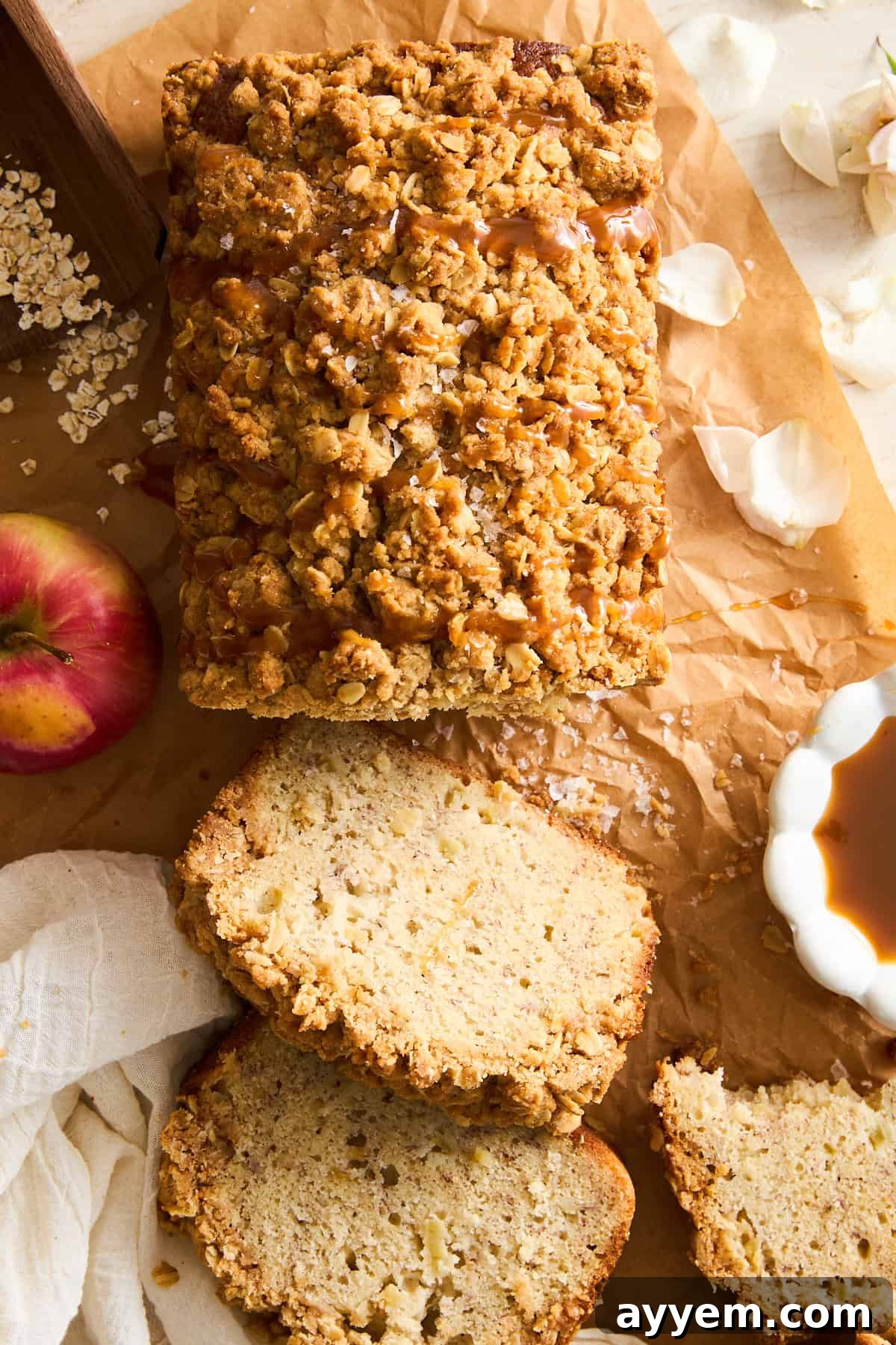 A loaf of crumbly apple banana bread with a streusel topping sits on brown parchment paper. Three slices are cut, and an apple, oats, a small bowl of caramel, and flower petals are nearby, evoking a warm, autumnal feel.