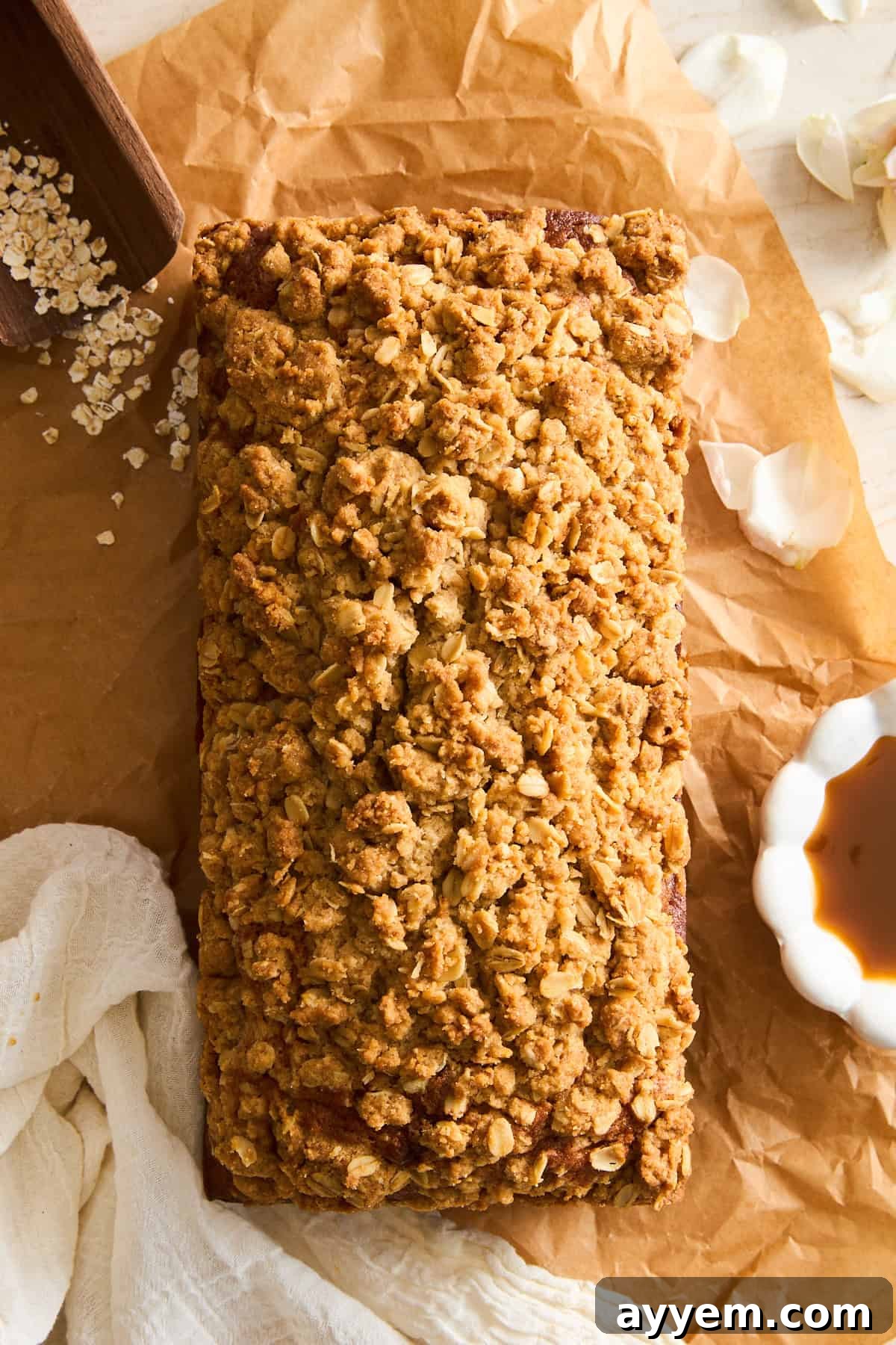 A loaf of apple banana bread with a golden oat crumble topping sits on brown parchment paper, surrounded by scattered oats, a white linen cloth, delicate flower petals, and a small bowl of golden syrup, creating an inviting, rustic scene.