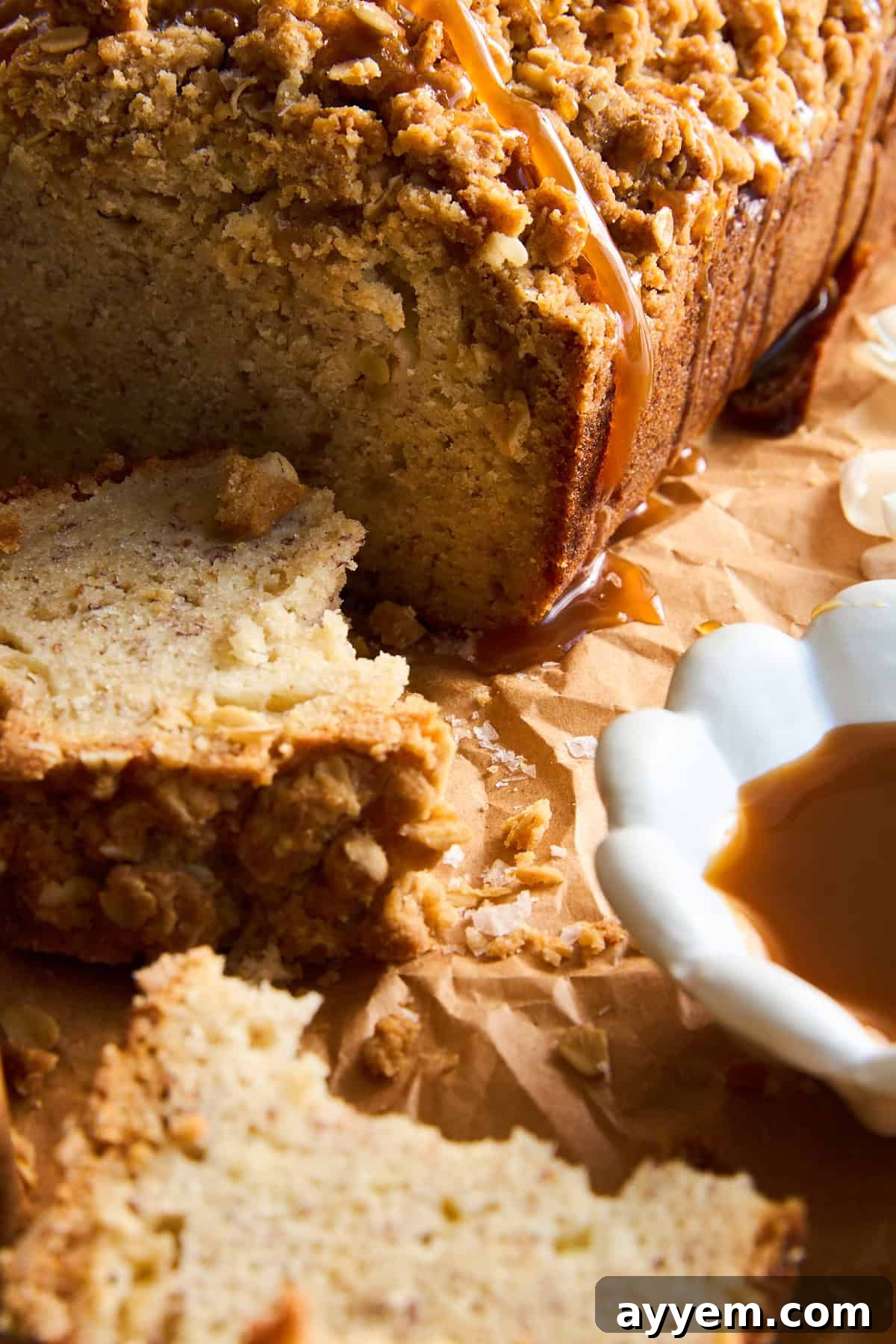 Close-up of sliced apple banana bread with a streusel topping and caramel drizzle, resting on brown parchment paper. A white dish with caramel sauce sits nearby, along with scattered crumbs, highlighting the rich texture.
