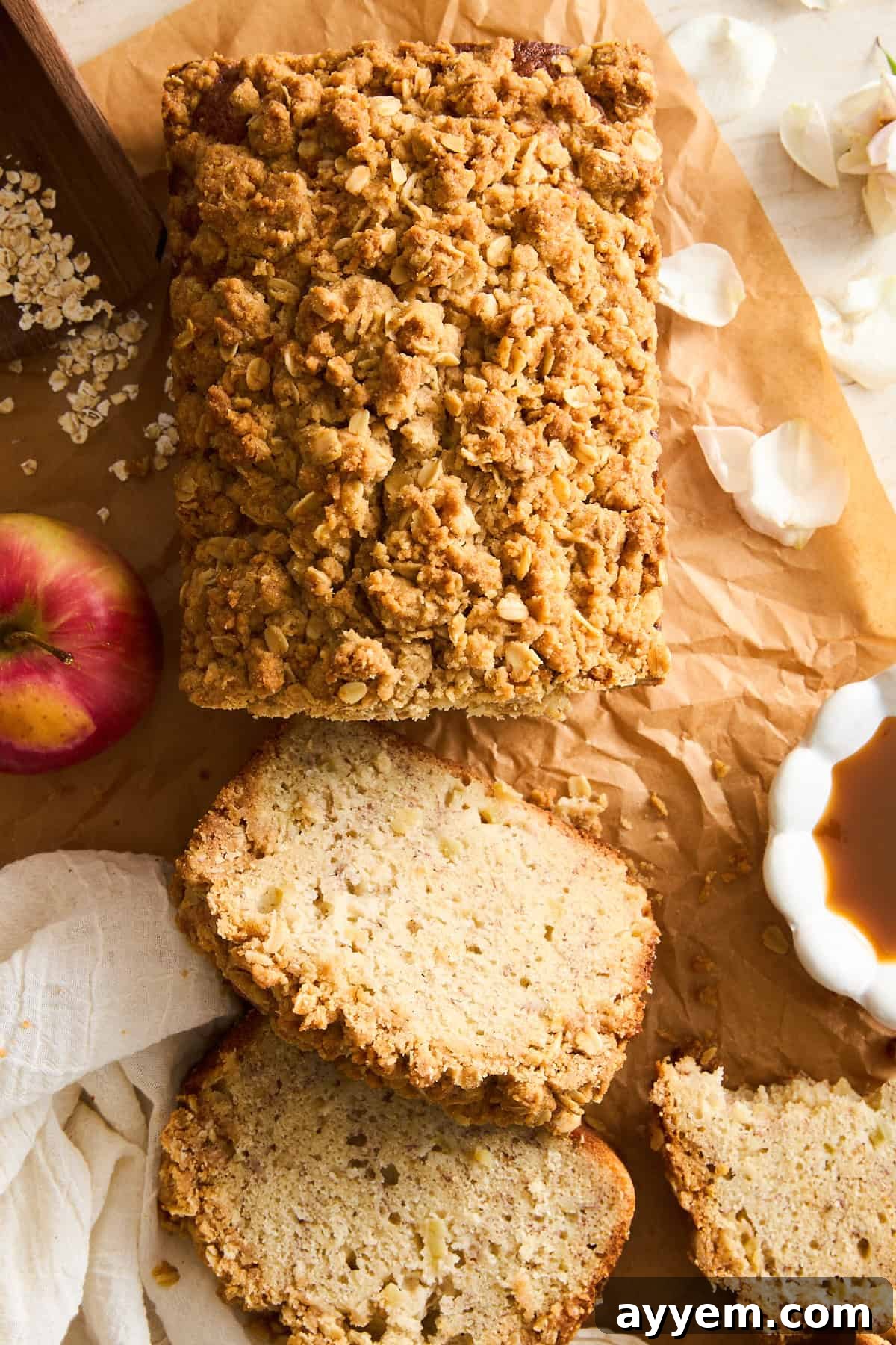 A loaf of apple crumble bread on parchment paper, partially sliced with three pieces cut. An apple, a white cloth, a bowl of caramel sauce, oats, and flower petals are placed around the bread, creating a cozy autumn scene.