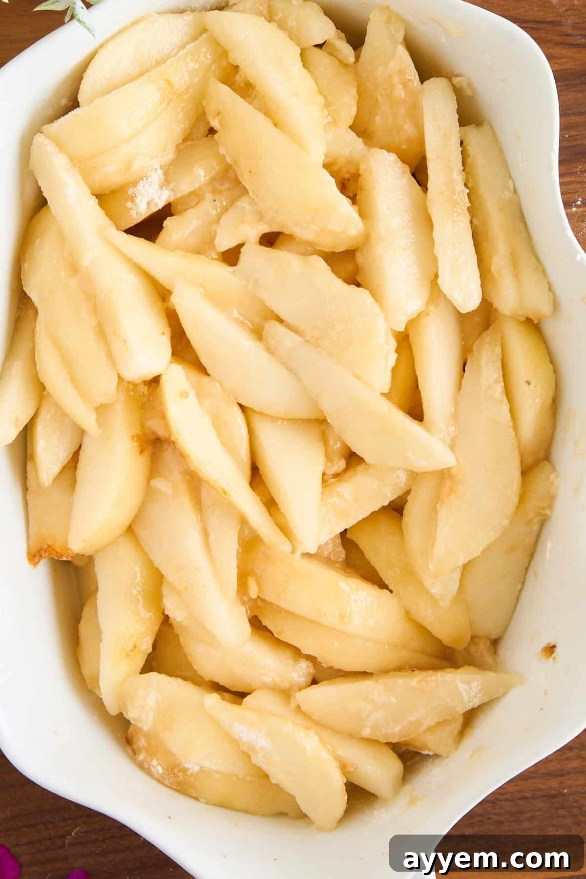 Sliced fresh pears coated with sugar and arranged in a white ceramic baking dish, ready for baking. The pears have a glossy appearance and are evenly distributed in the dish.