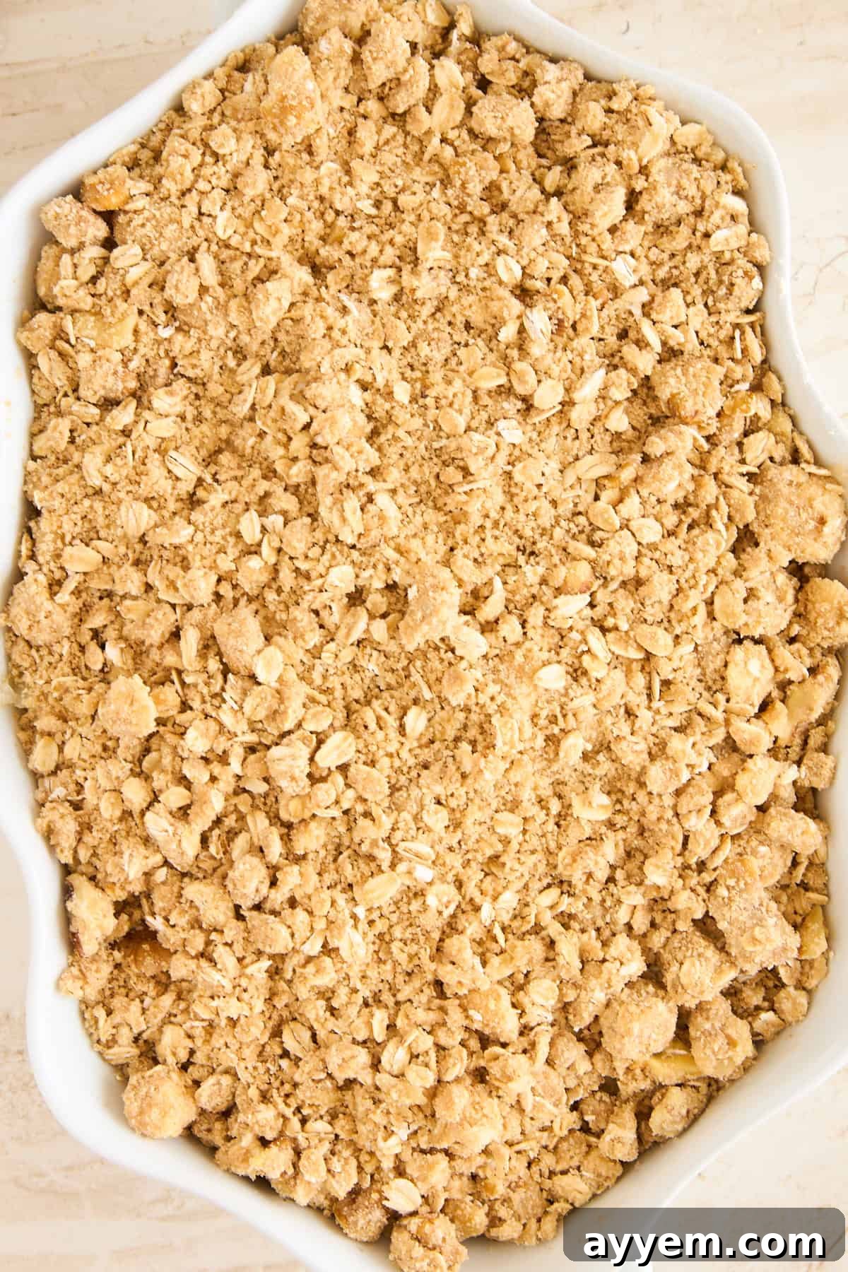 A close-up of an unbaked oat crumble topping in a white baking dish, showing a coarse mixture of oats, flour, butter, and sugar.