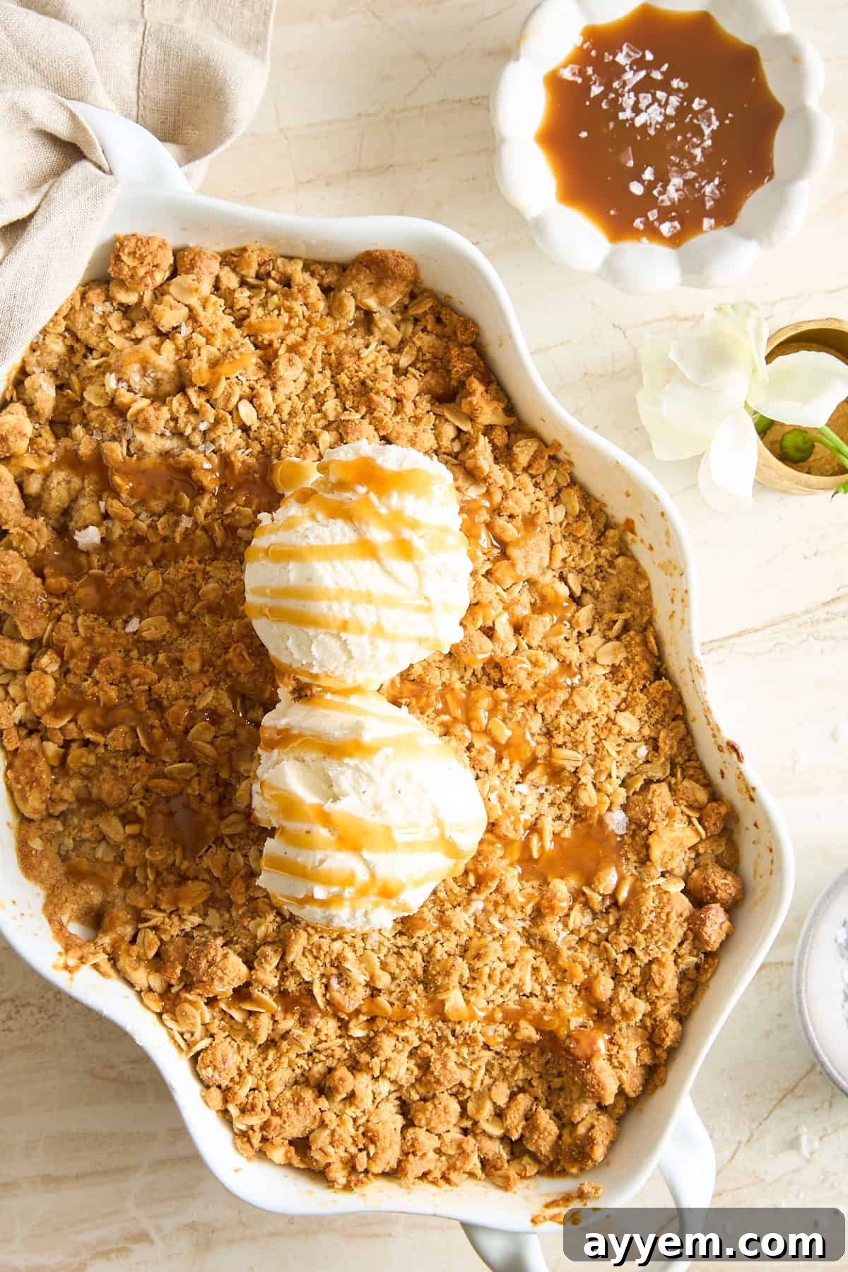 A baked pear crisp topped with two scoops of vanilla ice cream and caramel sauce in a white dish. Next to it is a bowl of caramel sauce sprinkled with sea salt and a white flower on a marble surface.