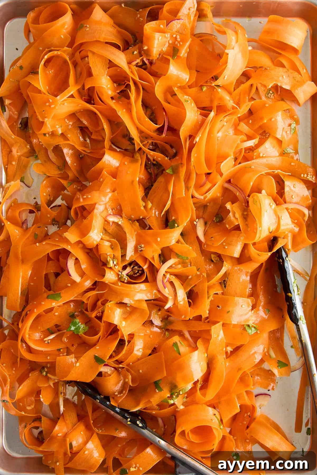A tray filled with thin ribbons of carrot salad, garnished with herbs and seeds, with metal tongs resting on the side.