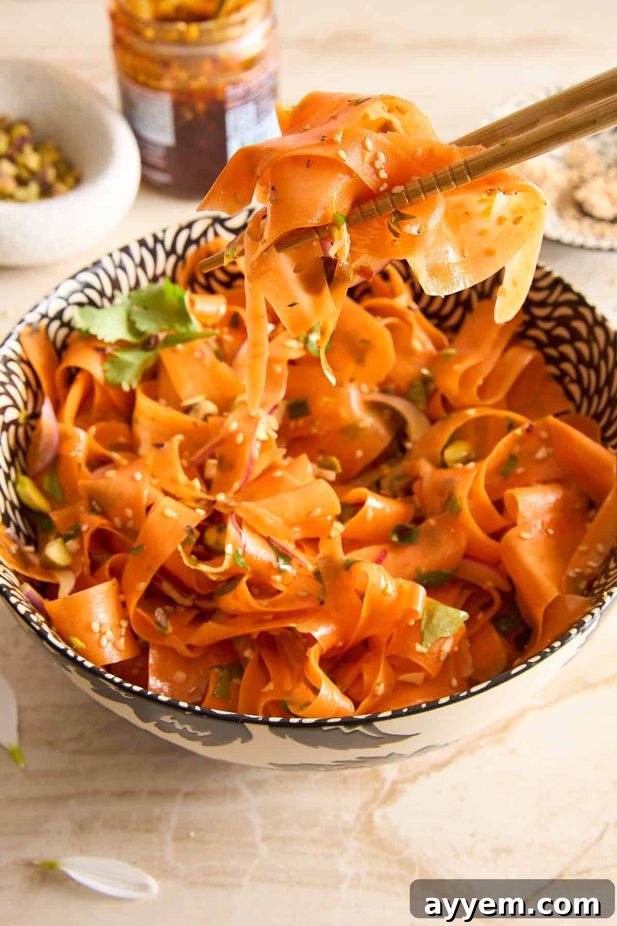 A patterned bowl filled with carrot ribbons garnished with herbs and seeds. Chopsticks hold up some carrot ribbons above the bowl. A jar of sauce and a small dish are visible in the background.