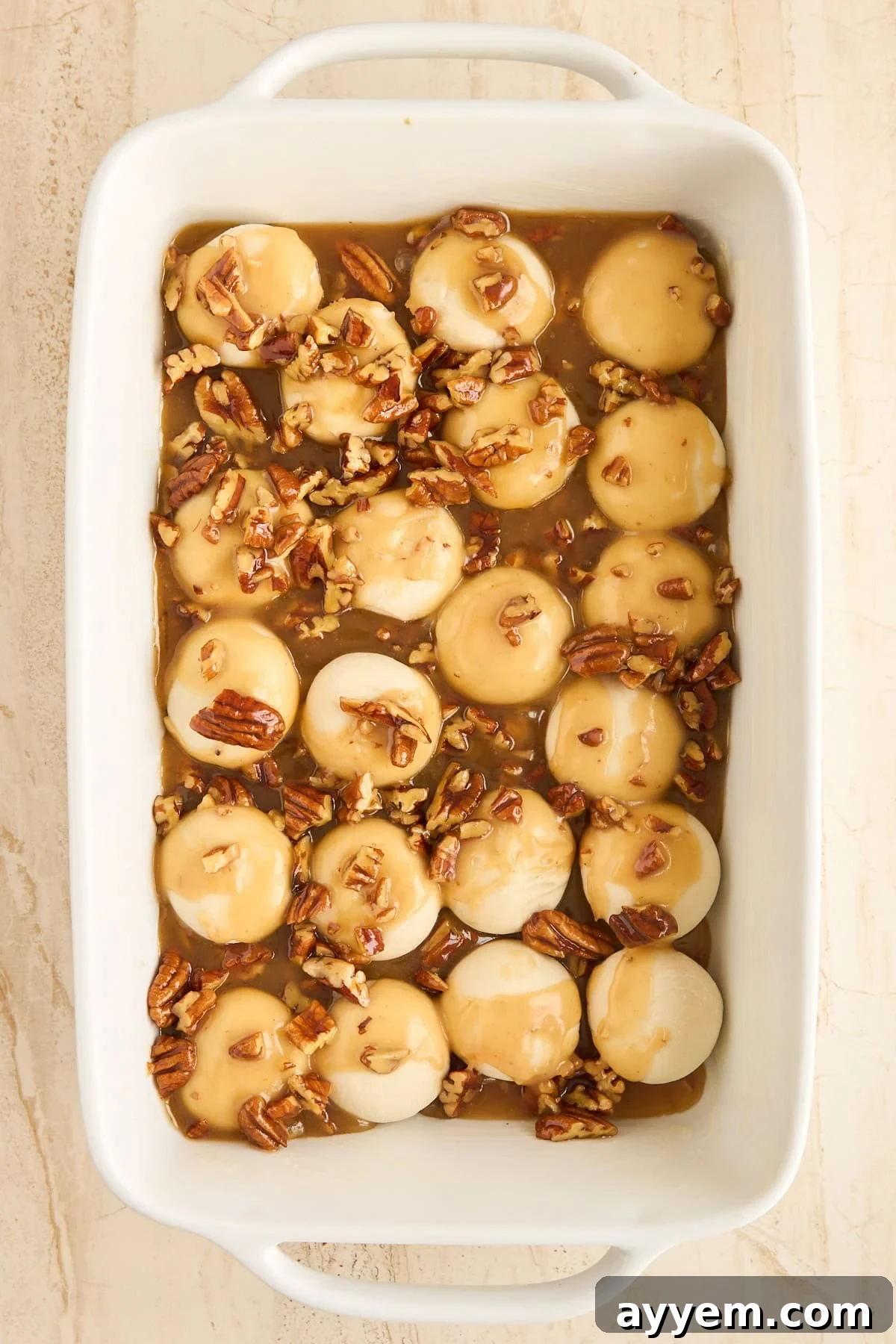 A white baking dish filled with unbaked, round dough balls topped with golden-brown caramel sauce and chopped pecans, ready for rising.