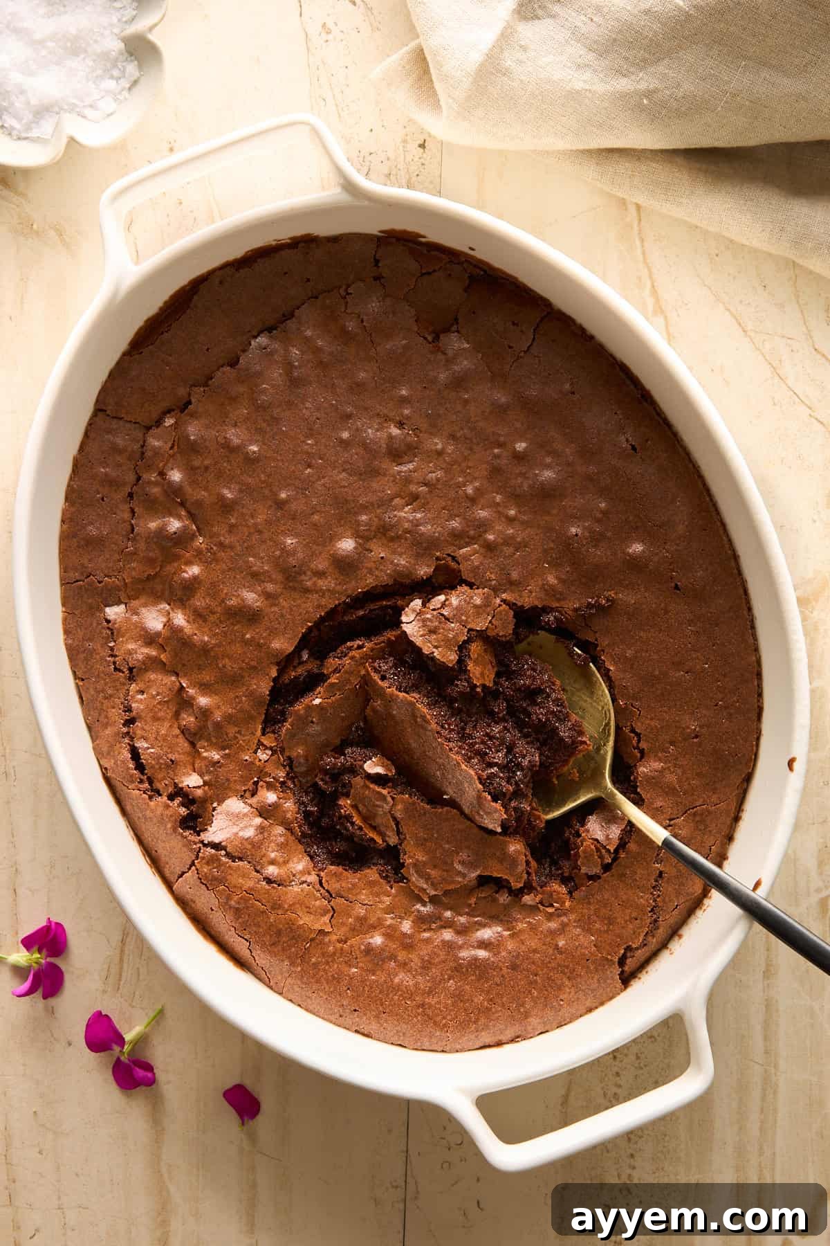 Oval white baking dish with a cracked, rich, baked chocolate dessert. A spoon is scooping out a portion from the center, revealing a gooey, luscious interior. Purple flower petals and a bowl of white powder are beside the dish on a light surface, suggesting a decadent treat.