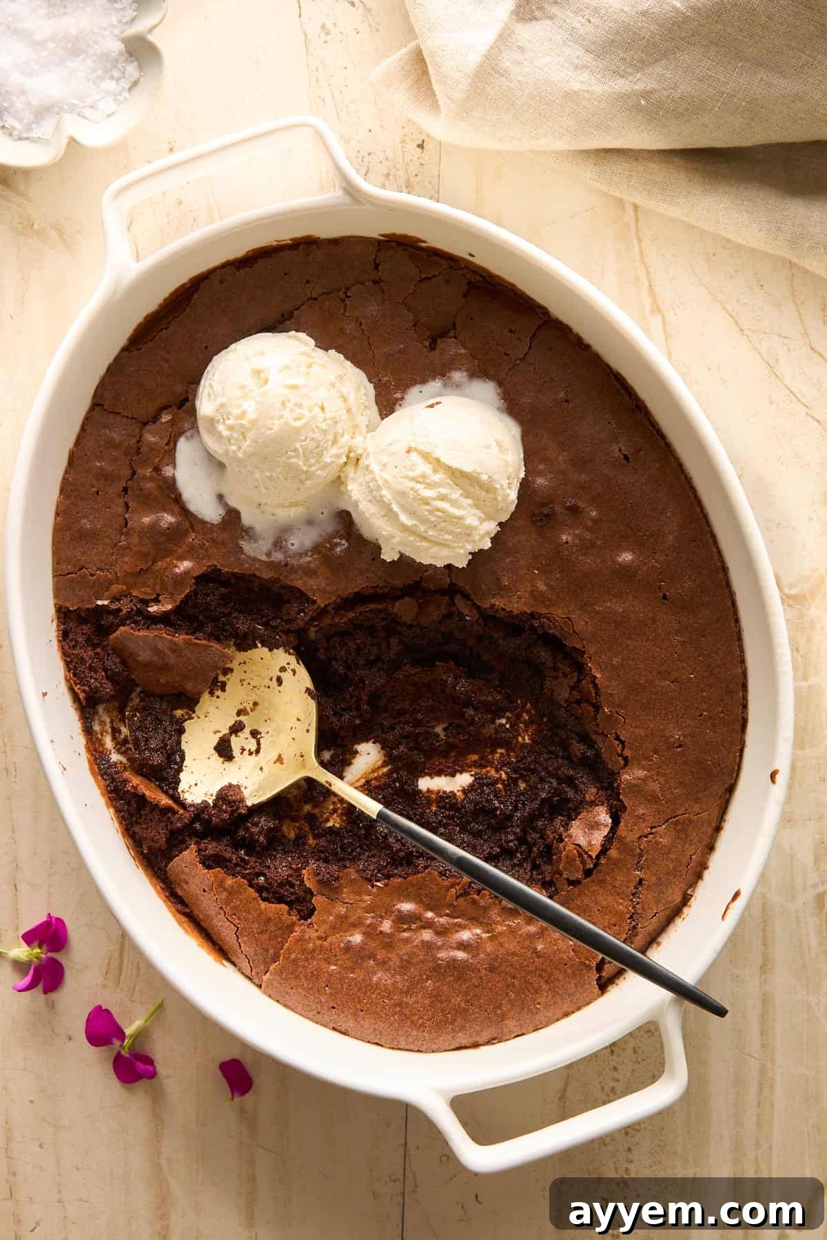 An oval white baking dish holds a chocolate cake with a large scoop missing, revealing a gooey interior. Two scoops of vanilla ice cream sit on top, with a serving spoon resting inside. Pink flowers are beside the dish, making for a picturesque dessert display.