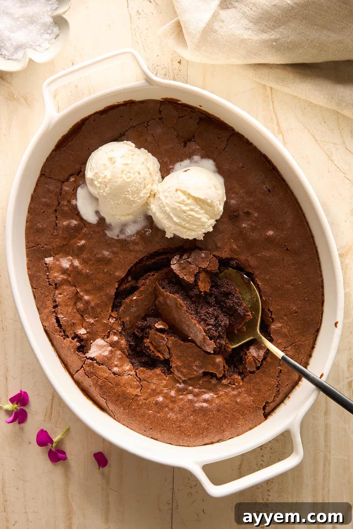 A white baking dish with chocolate brownie and two scoops of vanilla ice cream on top. A spoon has taken a serving out, showing the gooey brownie inside. Pink flowers and a beige cloth are nearby on a light surface.