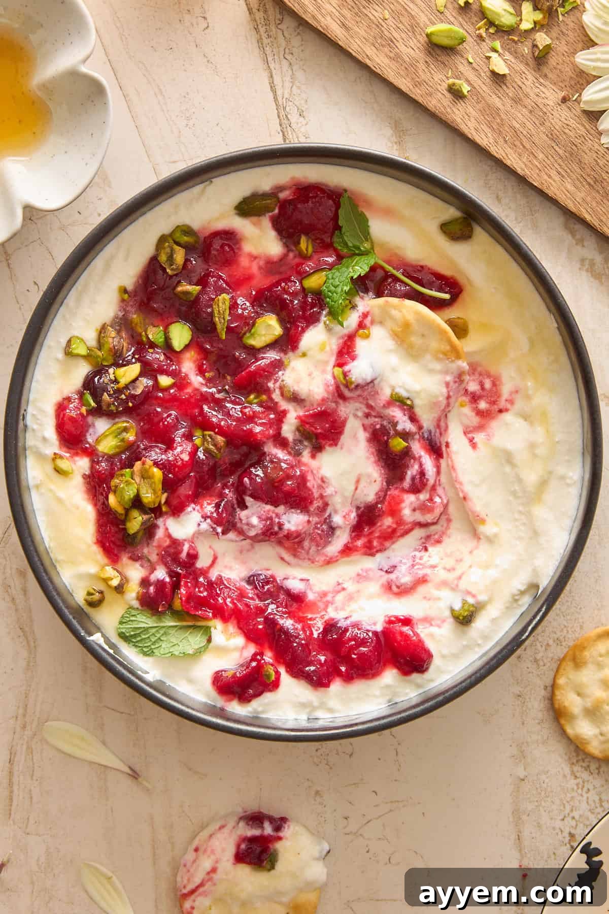 A close-up shot of the finished whipped feta dip. The creamy white feta base is topped with a rich, glistening swirl of red cranberry compote, adorned with bright green chopped pistachios and delicate fresh mint leaves. The dish rests on a light-colored surface, with scattered pistachios, mint sprigs, and a golden drizzle of honey visible around it, creating an inviting and appetizing scene.