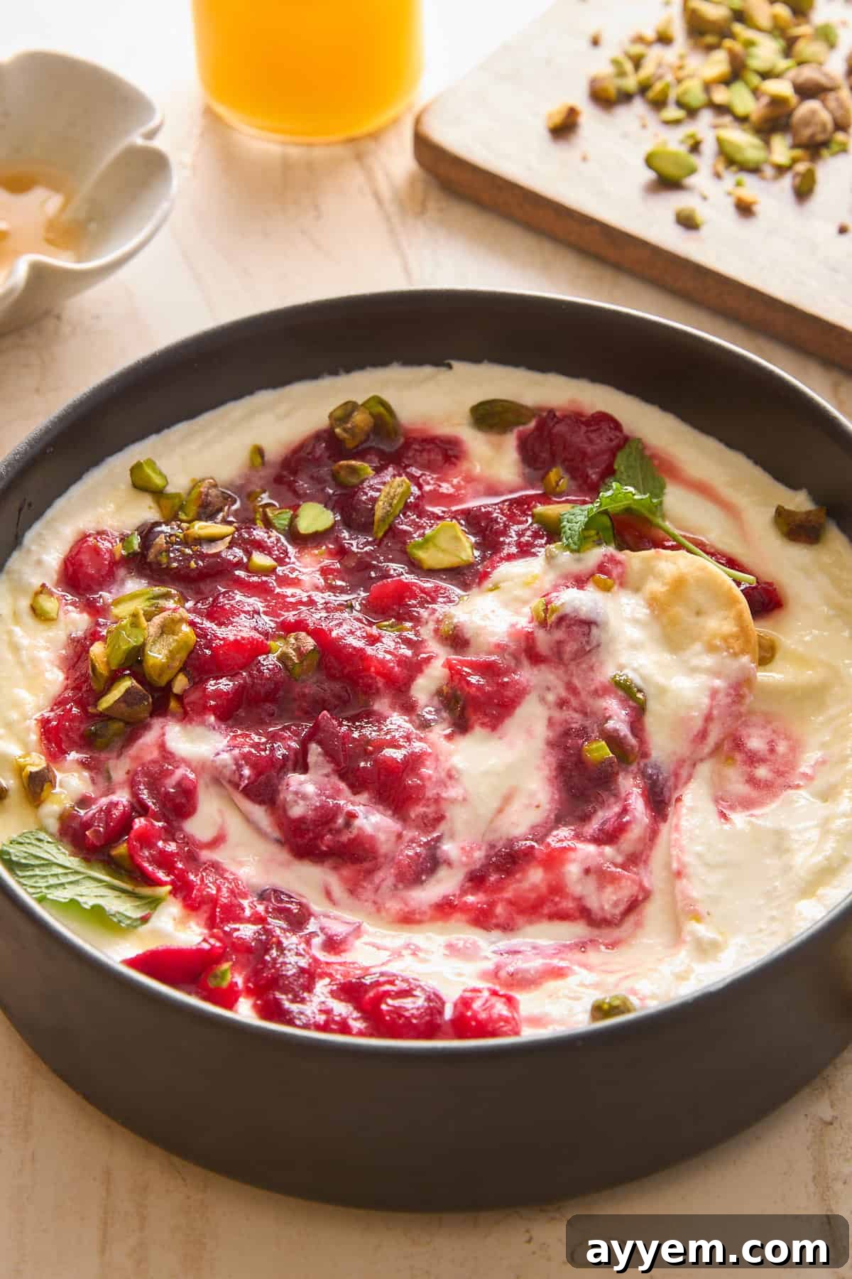 A bowl of creamy yogurt topped with swirling raspberry sauce, chopped pistachios, and fresh mint leaves sits on a light surface, with honey and more pistachios visible in the background.