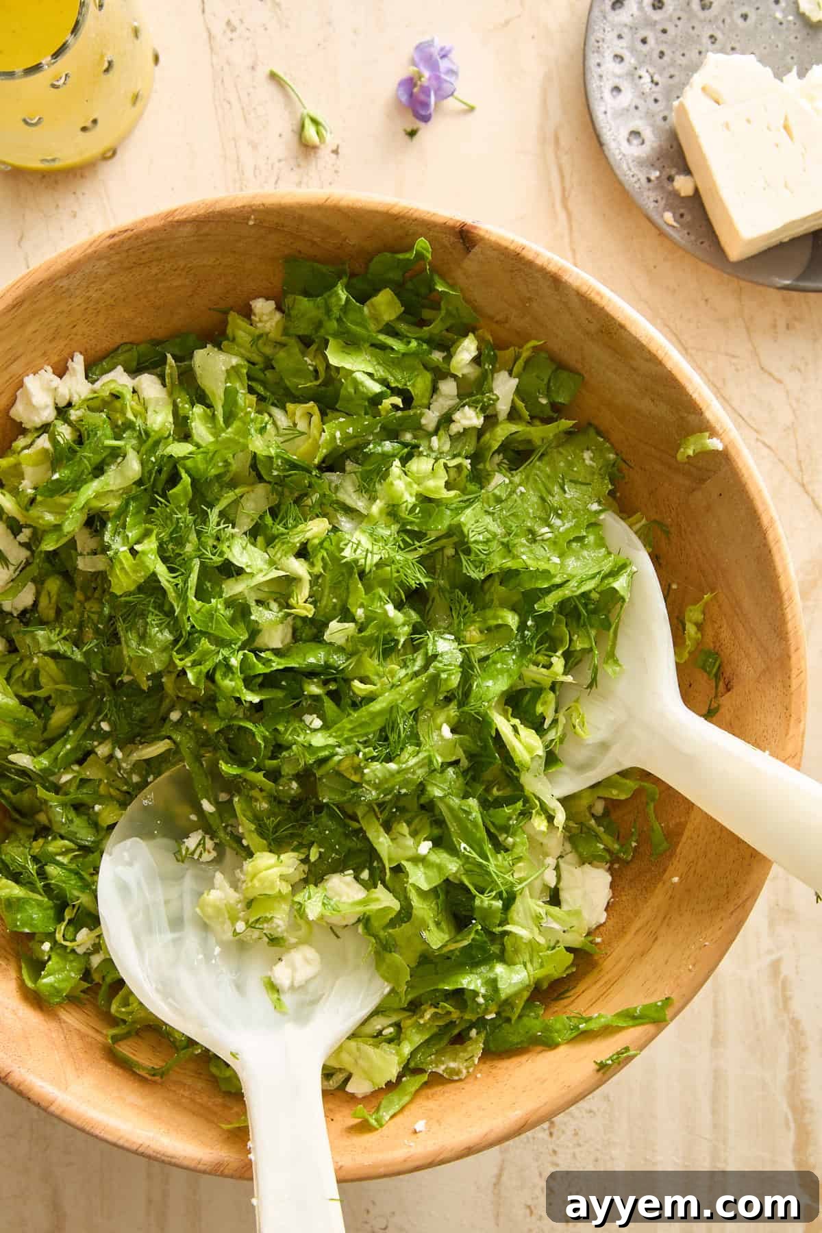 A wooden bowl filled with a fresh Maroulosalata, showcasing chopped green lettuce, generous feta cheese crumbles, and hints of dill. Two elegant white salad servers rest in the bowl, ready for serving. Adjacent to the bowl, on a light marble surface, are a plate with a block of feta cheese and a refreshing drink in a glass, completing a light meal setting.