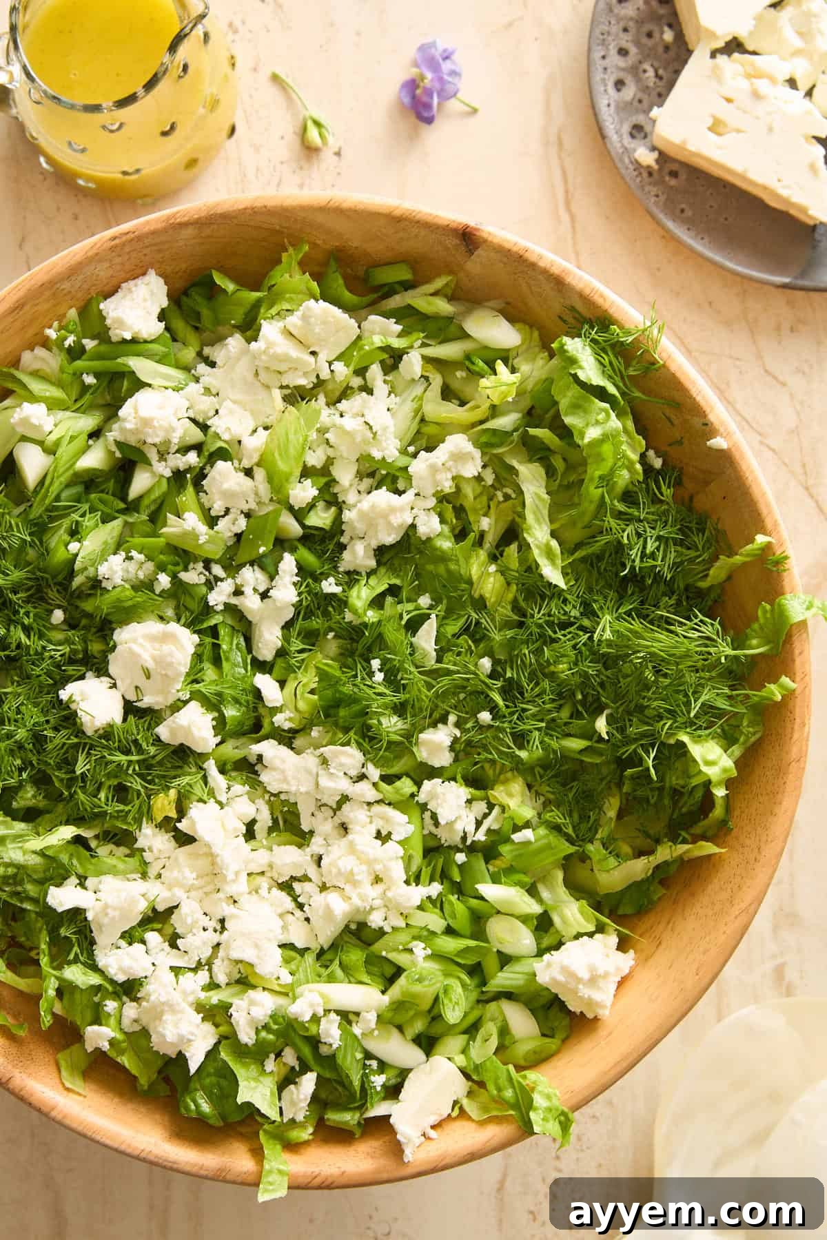 A vibrant wooden bowl brimming with chopped green lettuce, crisp cucumber slices, delicate fresh dill, and creamy crumbled feta cheese. Adjacent to the bowl, on a light surface, sits a glass container of bright salad dressing, a small plate with more feta, and a decorative purple flower, creating an inviting culinary scene.
