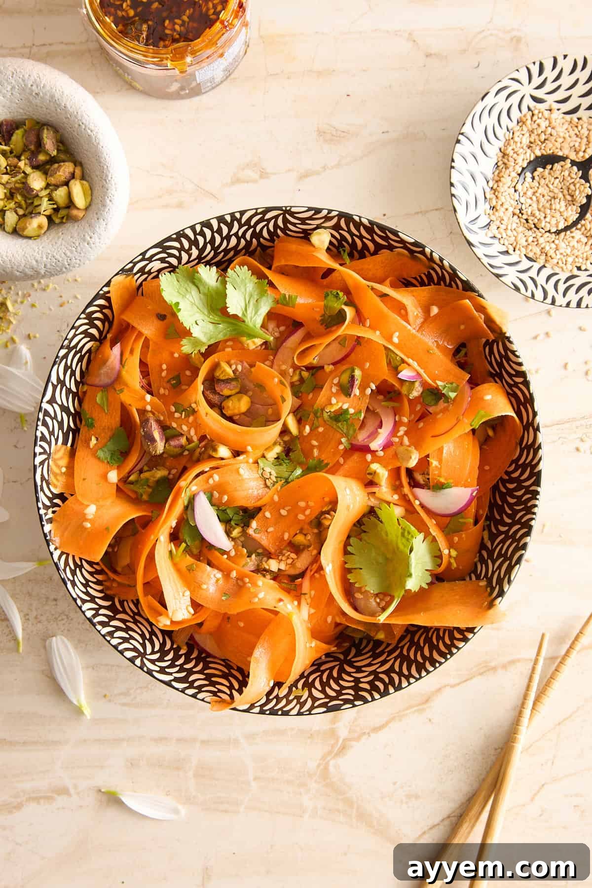 A bowl of carrot ribbon salad garnished with cilantro, red onion, sesame seeds, and pistachios sits on a light surface surrounded by a dish of sesame, a jar of chili flakes, and chopsticks. Dressed with vinaigrette.