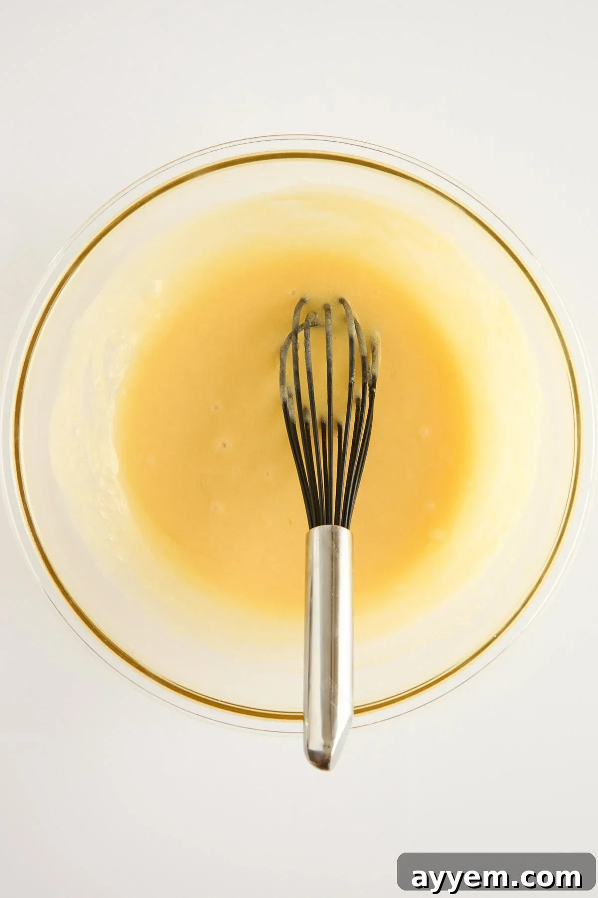 A metal whisk in a glass bowl containing pale yellow batter or mixture, set against a white background.