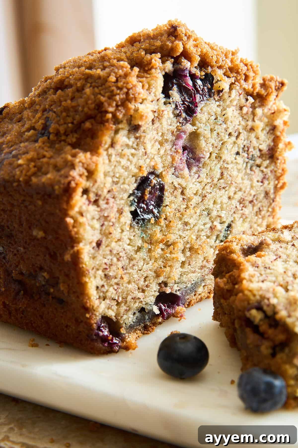 A close-up of a sliced blueberry bread loaf with a crumbly topping, showing moist texture and visible blueberries inside. Two fresh blueberries are placed on the marble surface nearby.