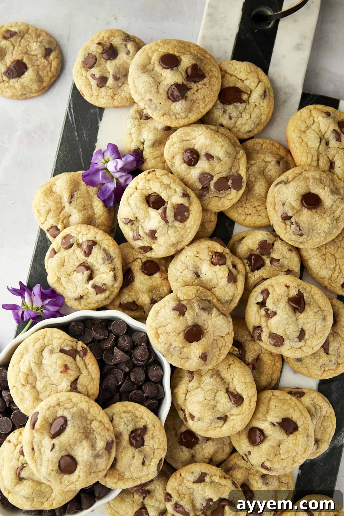 A pile of chocolate chip cookies is spread on a marble and wood board with purple flowers and a small bowl of chocolate chips beside them.
