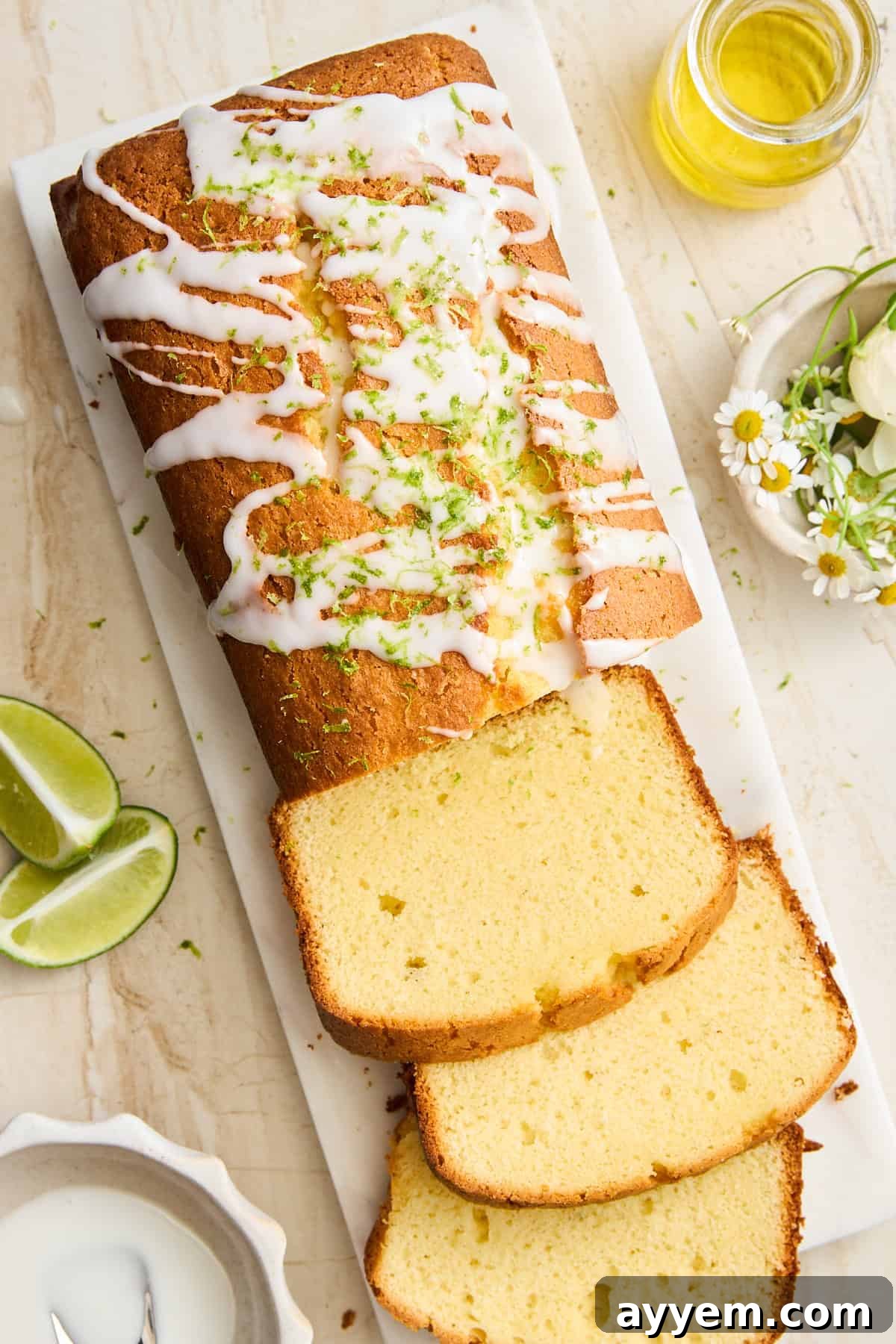 A loaf of glazed pound cake, drizzled with white icing and topped with lime zest, sits on a marble board. Several slices are cut, with lime wedges and a small jar of oil nearby, and flowers as decoration.