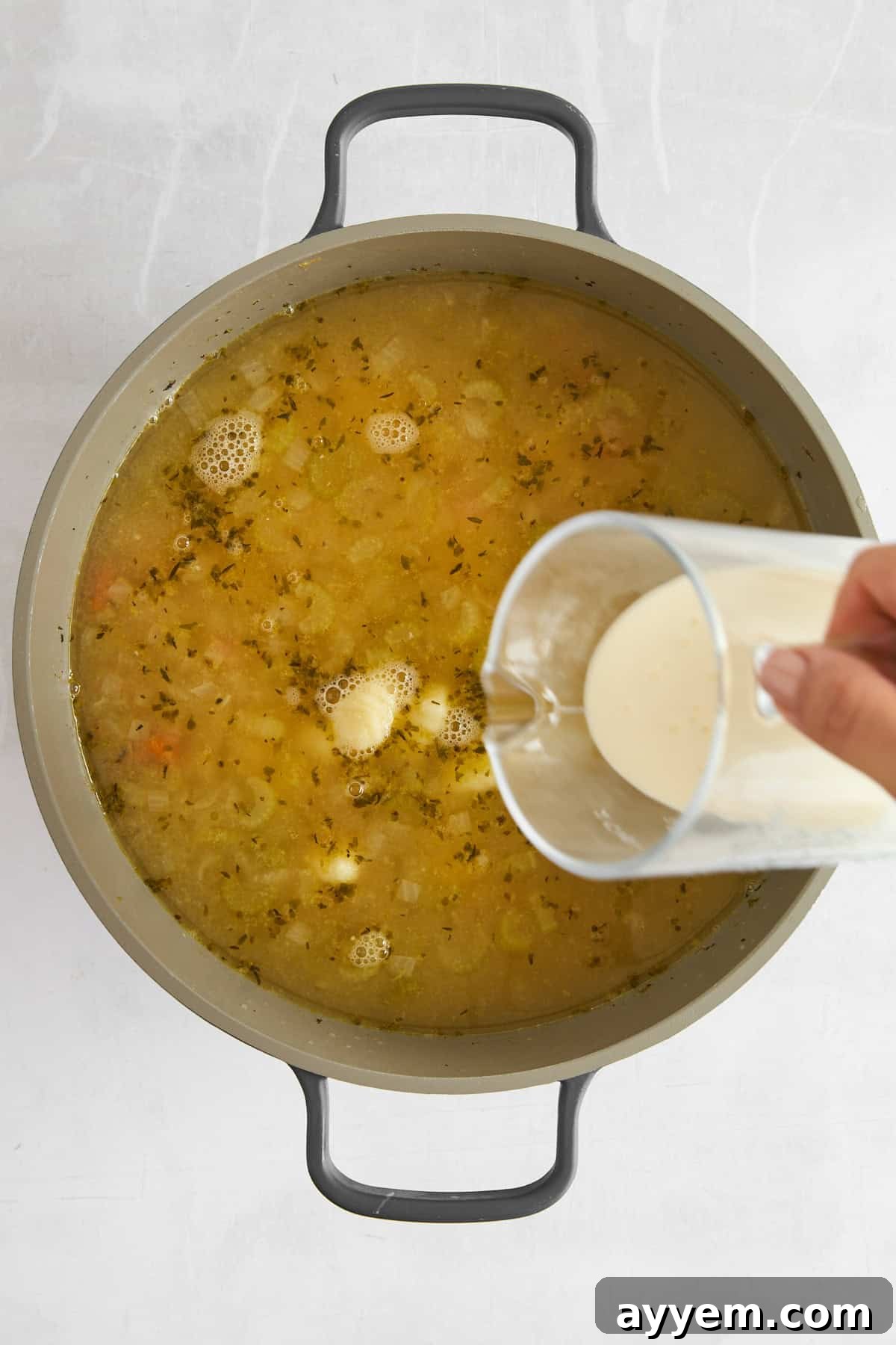 A hand is pouring a creamy liquid from a measuring cup into a pot of soup with herbs and vegetables on a white surface.