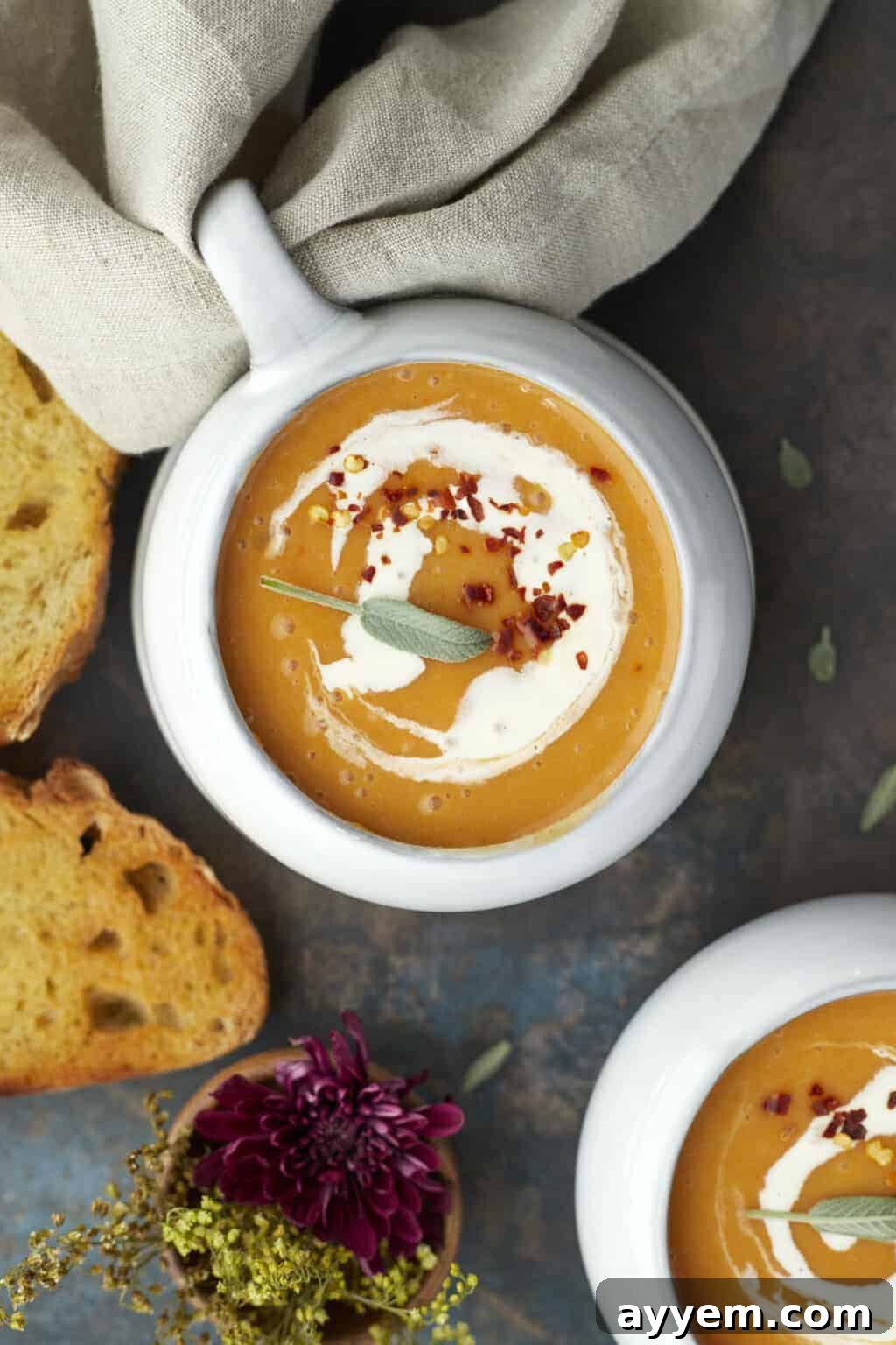 A bowl of creamy orange soup garnished with a swirl of cream, crumbled red pepper, and a sage leaf, with slices of toasted bread and a purple flower nearby.