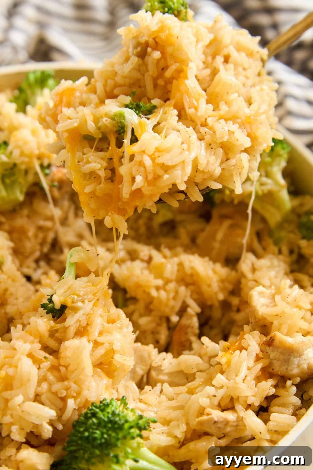 Close-up of a spoon lifting cheesy rice with broccoli out of a dish, with melted cheese stretching from the spoonful. Rice and broccoli are mixed together, and cheese is visible throughout the dish.
