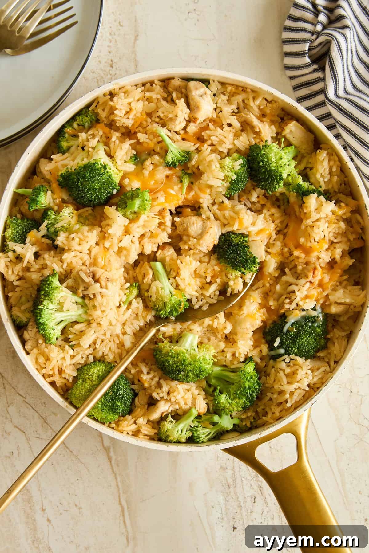 A skillet filled with rice, broccoli florets, and pieces of chicken, with a gold serving spoon resting inside. A striped cloth and stacked plates with forks are nearby on a light surface.